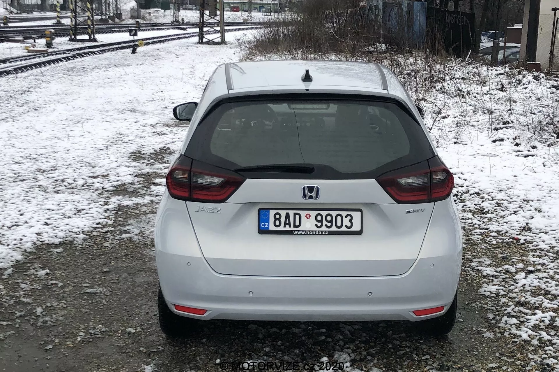 La imagen muestra la vista trasera de un Honda Jazz 2020 blanco, estacionado sobre un suelo nevado cerca de las vías del tren bajo condiciones de luz difusa o nublada. El coche ocupa la mayor parte del marco, con una vista trasera centrada que muestra su matrícula de estilo europeo, limpiaparabrisas trasero, luz de freno de montaje alto y el emblema de Honda. La insignia del modelo 'JAZZ' es visible en la parte inferior izquierda del portón trasero, y hay una cámara de visión trasera sobre la matrícula. Las luces traseras envuelven parcialmente los lados, con reflectores en la parte inferior del parachoques, que también tiene una sección negra integrada. Las señales de suciedad sugieren que el coche ha sido conducido por carreteras mojadas o fangosas. La nieve y los árboles desnudos indican que es invierno o principios de primavera.