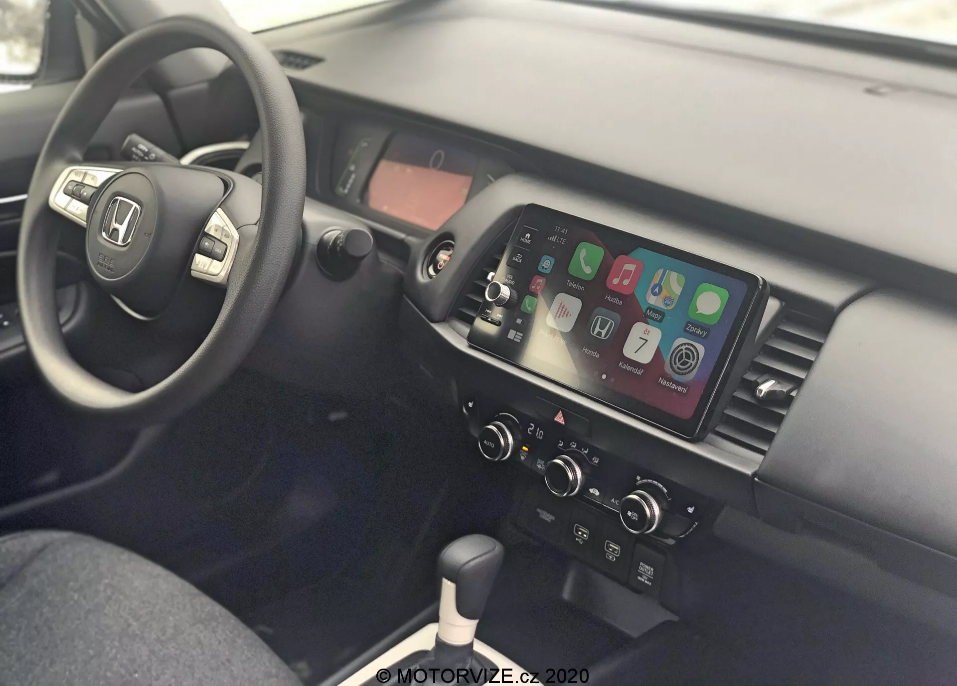 Vista interior de un Honda Jazz 2020, mostrando el área del conductor y la consola central desde el lado del pasajero. El volante está a la izquierda y cuenta con controles montados para audio, conectividad telefónica y control de crucero, lo que indica un diseño para conducir por la derecha. La consola central cuenta con una pantalla táctil que muestra la integración de smartphones como Apple CarPlay o Android Auto, controles manuales del clima, palanca de cambios para transmisión manual, puertos USB y una toma de corriente de 12V, enfatizando la conectividad moderna y el diseño funcional.