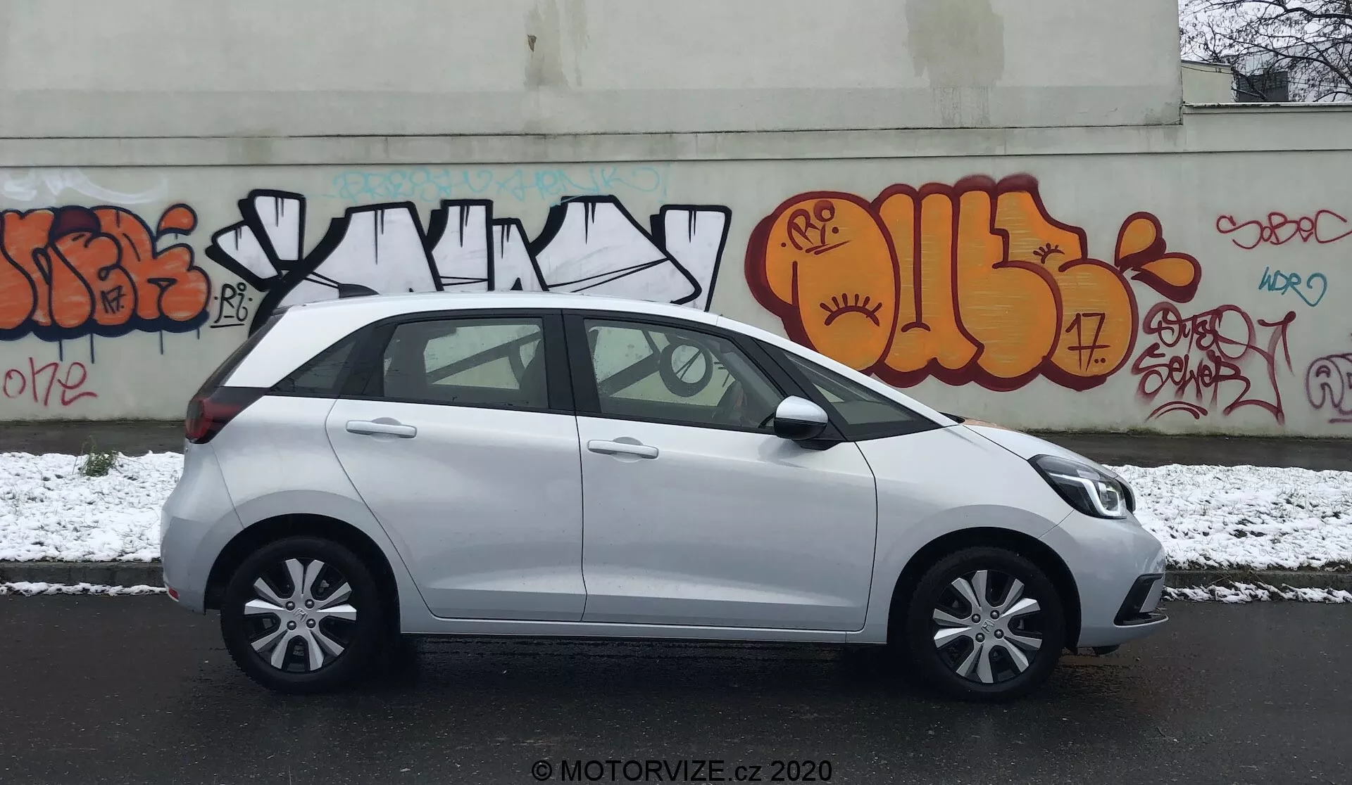 Perfil lateral de un Honda Jazz compacto y blanco del 2020 estacionado en una calle urbana con grafitis en la pared de fondo. La escena es en un día nublado con una ligera capa de nieve en el suelo, lo que sugiere condiciones invernales. La imagen captura el lado del conductor, mostrando manijas de las puertas del color del carro, espejos laterales, llantas de aleación de cinco radios y ventanas estándar con un pilar B negro. El vehículo parece limpio con algunas manchas húmedas de la nieve. Es visible la marca de agua 'MOTORVIZE.CZ 2020', indicando la fuente de la foto.