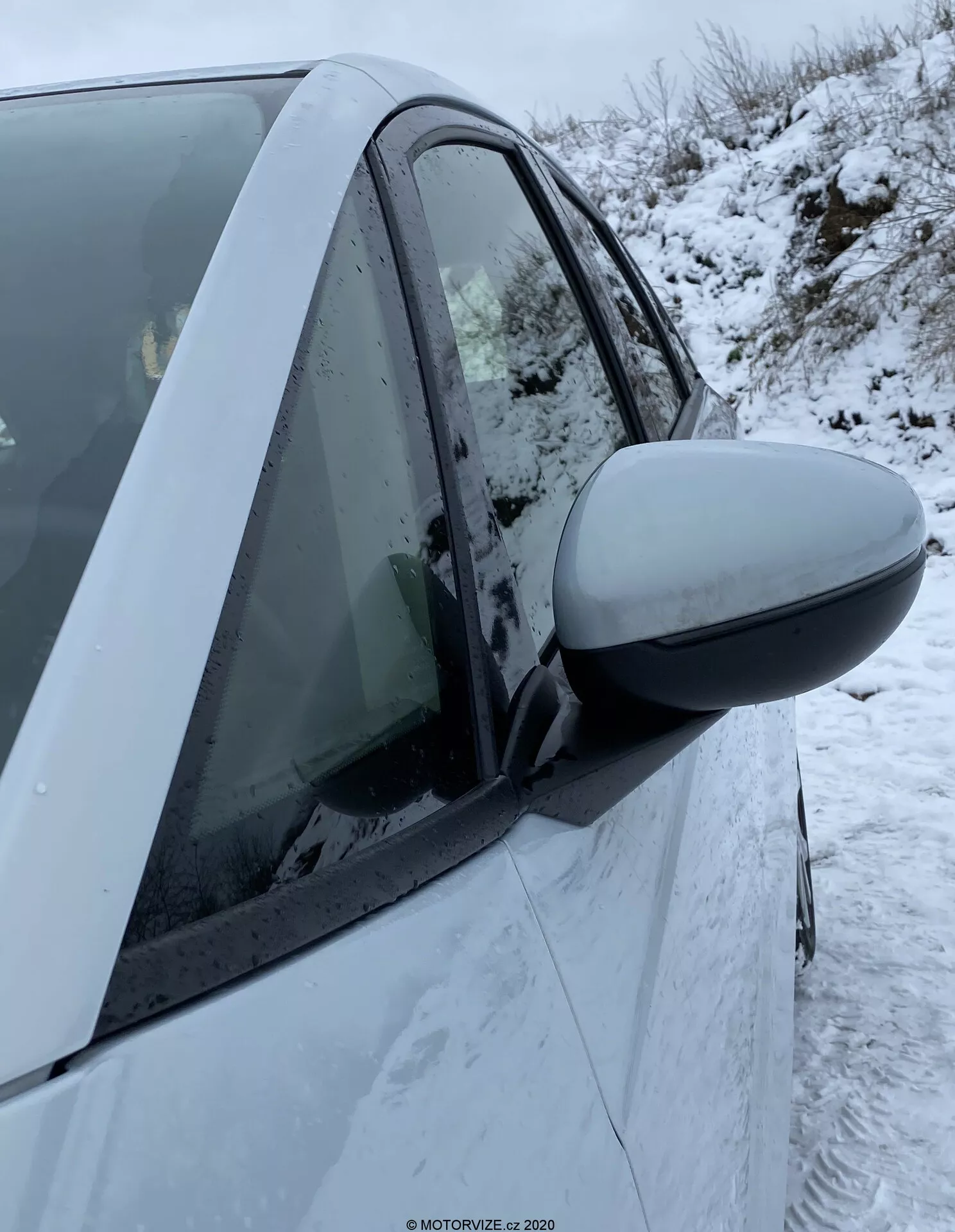 Primer plano del lado derecho de un Honda Jazz 2020 que muestra la zona de la puerta del conductor. El espejo lateral prominente se extiende desde la izquierda de la imagen hasta el centro, indicando uso reciente o próximo. La carcasa del espejo coincide con el color del coche, sugiriendo que es una característica estándar. Elementos de diseño moderno incluyen un espejo aerodinámico y la carrocería intacta. La ventana está subida con condensación en el interior y se pueden ver copos de nieve en la puerta y la ventana. El pilar B visible y partes de ambas puertas, delantera y trasera, indican un vehículo de cuatro puertas. El indicador lateral no es visible. El fondo muestra condiciones invernales con nieve en el suelo.