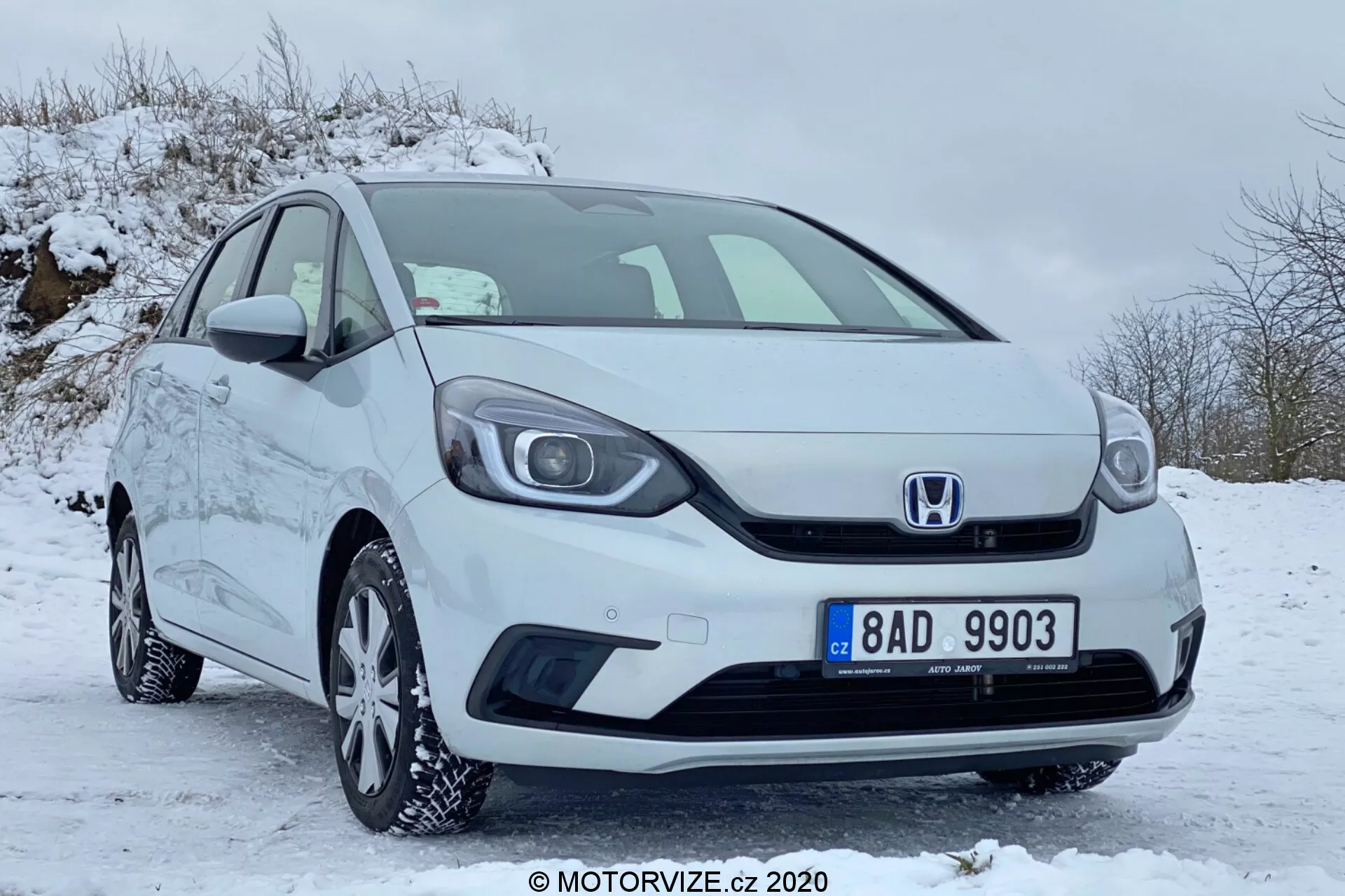 Vista de tres cuartos frontal de un coche compacto blanco Honda Jazz 2020 estacionado en un área cubierta de nieve, mostrando la parrilla con el emblema de Honda, el parachoques delantero, los faros, las llantas de aleación con neumáticos de invierno, los espejos laterales y las manijas de las puertas, todo en una condición bien mantenida y limpia.