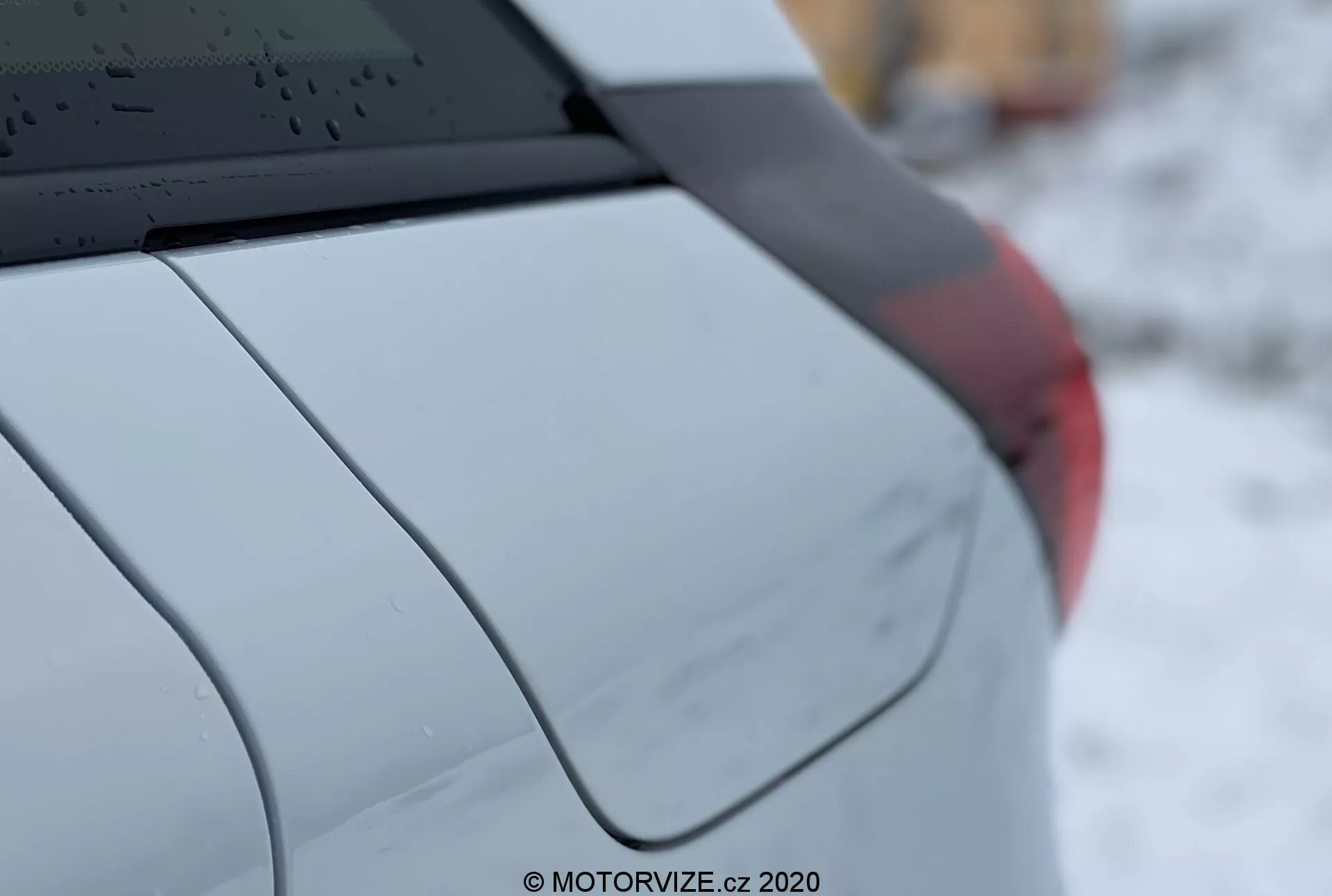 Detalle en primer plano del Honda Jazz 2020, mostrando el cuarto trasero derecho del coche desde una vista lateral de tres cuartos. Las características incluyen una ventana trasera con líneas de deshielo, una antena de aleta de tiburón en el techo, el pilar C acentuando el diseño del coche, un guardabarros trasero suavemente curvado, un fragmento de un conjunto de luces traseras y un acabado de pintura metálica o perlada. La poca profundidad de campo destaca los detalles del vehículo mientras el fondo está desenfocado. La marca de agua dice 'MOTORVIZE.cz, 2020.'