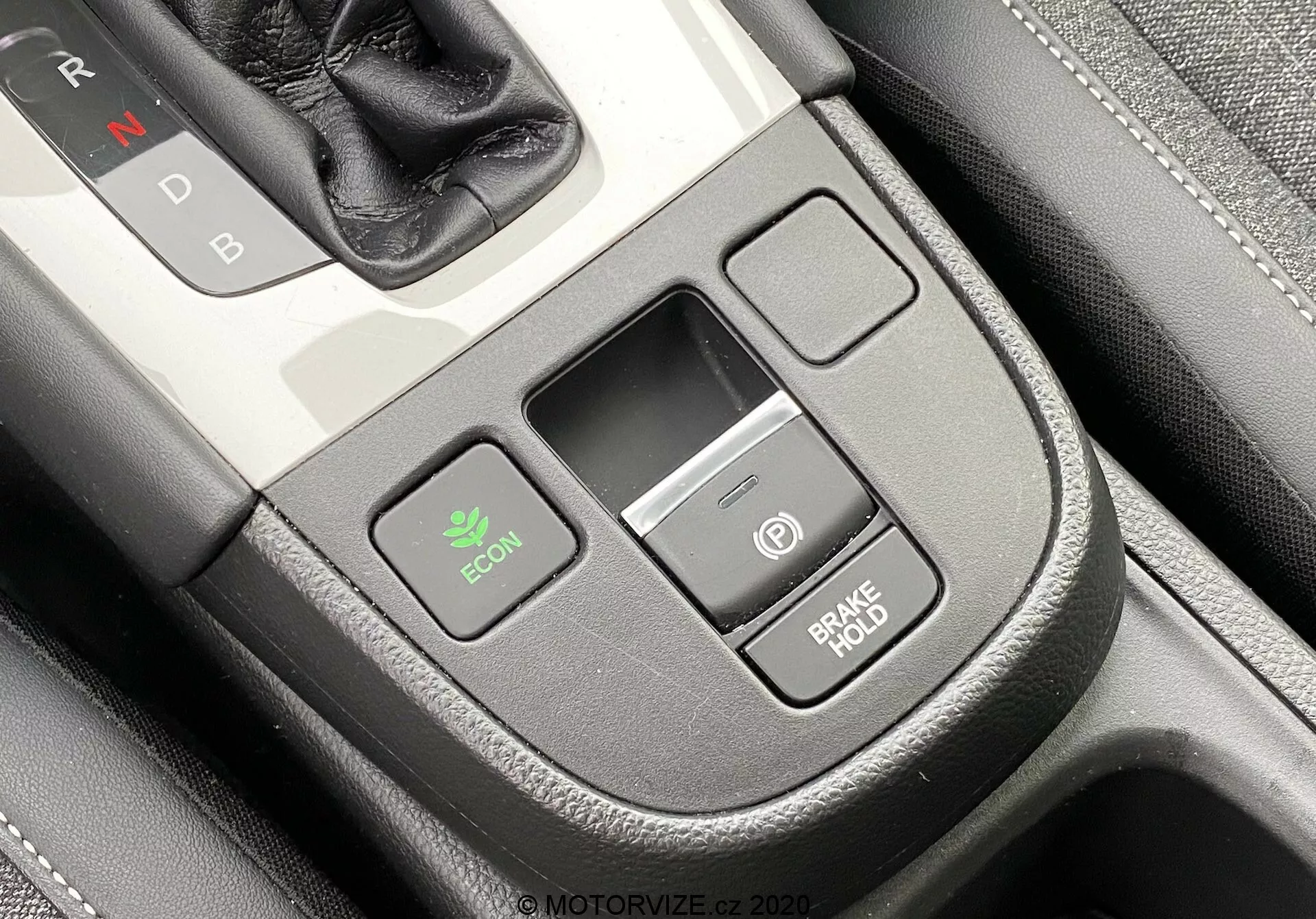 Primer plano de la consola central interior de un Honda Jazz 2020 desde una perspectiva ligeramente inclinada del conductor. El selector de cambios con 'N' de Neutral es parcialmente visible en la parte superior derecha, indicando transmisión automática. Destacan el botón ECON a la izquierda para el modo económico, una pequeña ranura de almacenamiento rectangular en el centro y dos botones apilados a la derecha—uno con un icono de encendido y el otro etiquetado 'BRAKE HOLD,' que evita el desplazamiento en paradas. La consola tiene un acabado premium con textura similar a cuero y detalle de costuras.