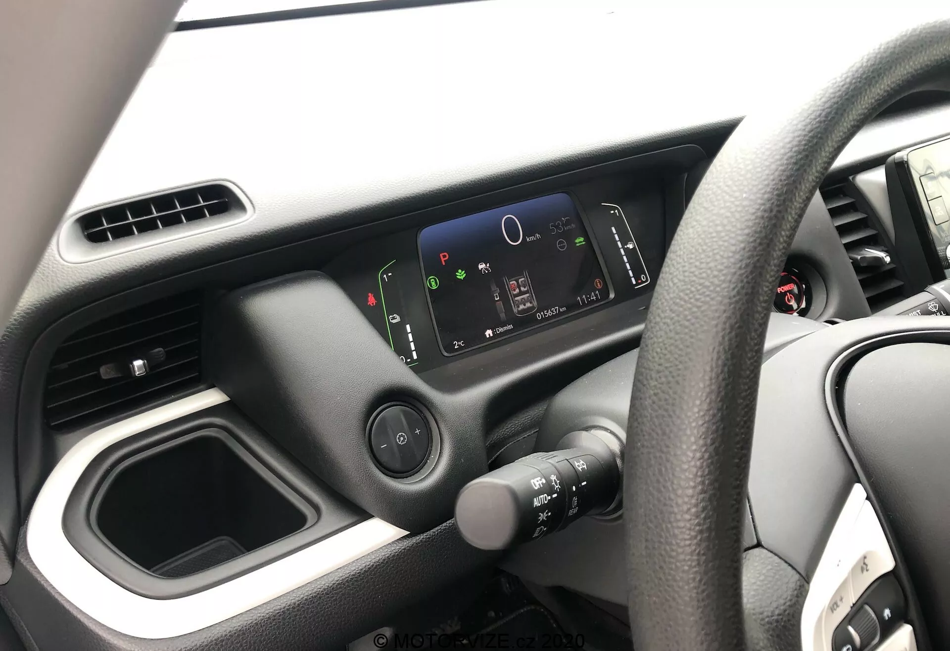 Vista del interior del Honda Jazz 2020, centrada en el lado del conductor desde la perspectiva del pasajero. La imagen muestra el volante en el lado derecho con un diseño de cuatro radios, el tablero de mando minimalista con un panel de instrumentos digital de alta resolución y el diseño simple de la consola central con un compartimento de almacenamiento vacío y salidas de aire. Solo se ve una parte de la puerta del conductor.