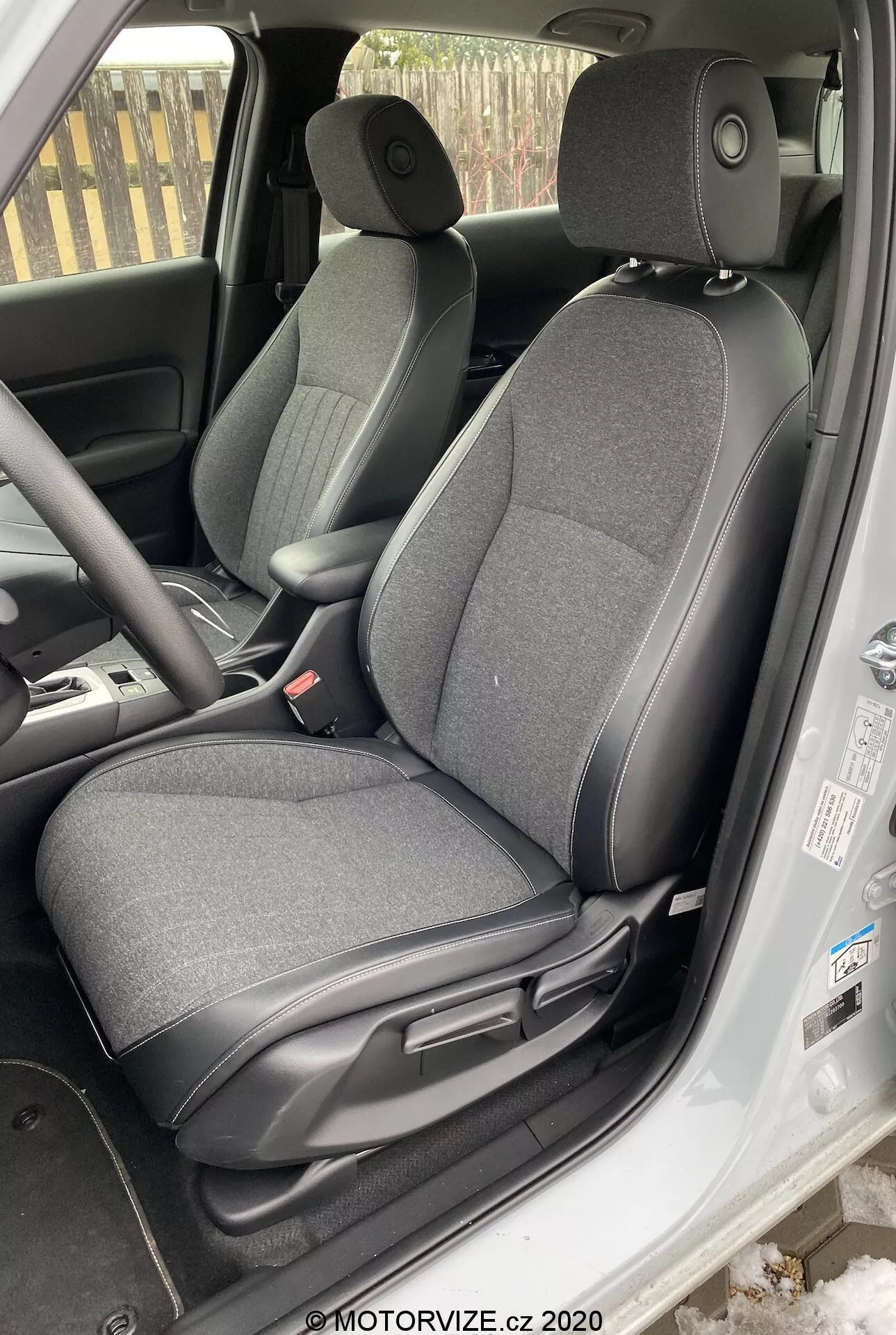 La imagen muestra el interior de un Honda Jazz 2020, centrado en el área del asiento del conductor tomada desde un ángulo ligeramente por encima del nivel ocular desde el exterior del vehículo. El asiento del conductor ajustable está tapizado en tela oscura. Son visibles las palancas de ajuste para deslizar y reclinar el asiento, el panel interior de la puerta con controles de ventanas eléctricas, el volante parcialmente visible con controles montados en el volante, una porción del tablero, la alfombrilla del piso y el área de los pedales que indican una posible transmisión manual, parte de la consola central y el cinturón de seguridad del conductor colgando al costado.