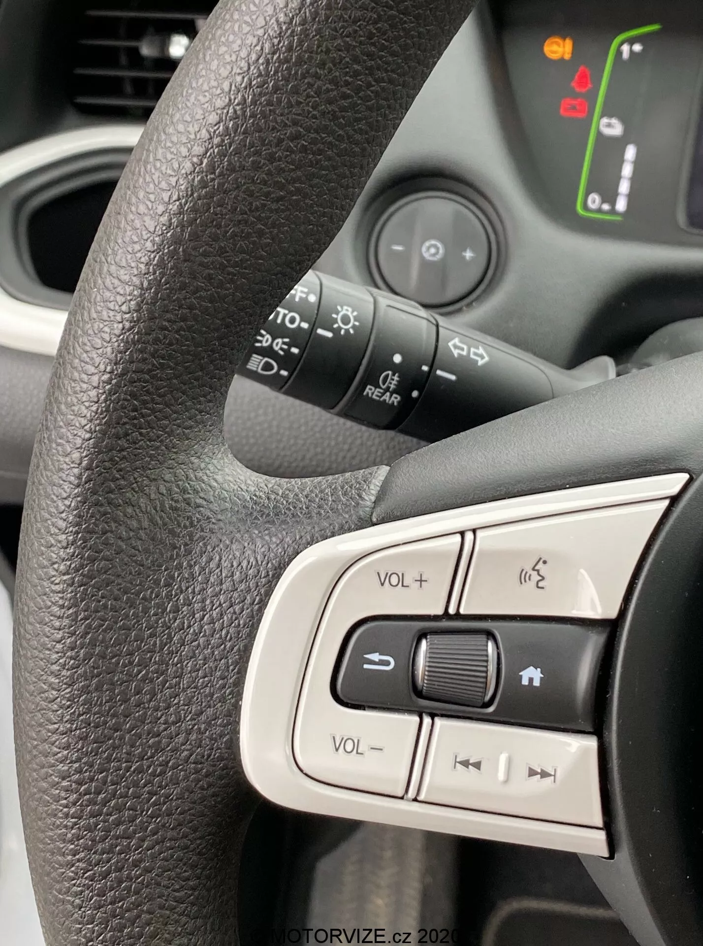 Vista de cerca del lado izquierdo del volante del Honda Jazz 2020 centrada en los botones de control integrados. Volante texturizado negro con agarre ergonómico, que muestra los controles de volumen, el botón de silencio y los botones de navegación de medios. Fondo ligeramente borroso revela símbolos iluminados en el tablero del coche, indicando que el vehículo está encendido. La luz natural brillante capta el diseño moderno y fácil de usar, enfatizando la ergonomía interior y las características tecnológicas.