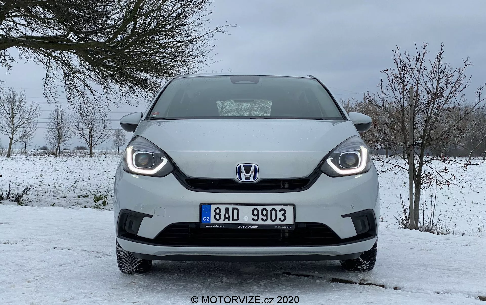 Vista frontal de un Honda Jazz 2020 en condiciones invernales, mostrando un diseño de parrilla distintivo, faros visibles con luces diurnas LED y placa de matrícula central, estacionado en una carretera cubierta de nieve con cielo nublado y árboles sin hojas.