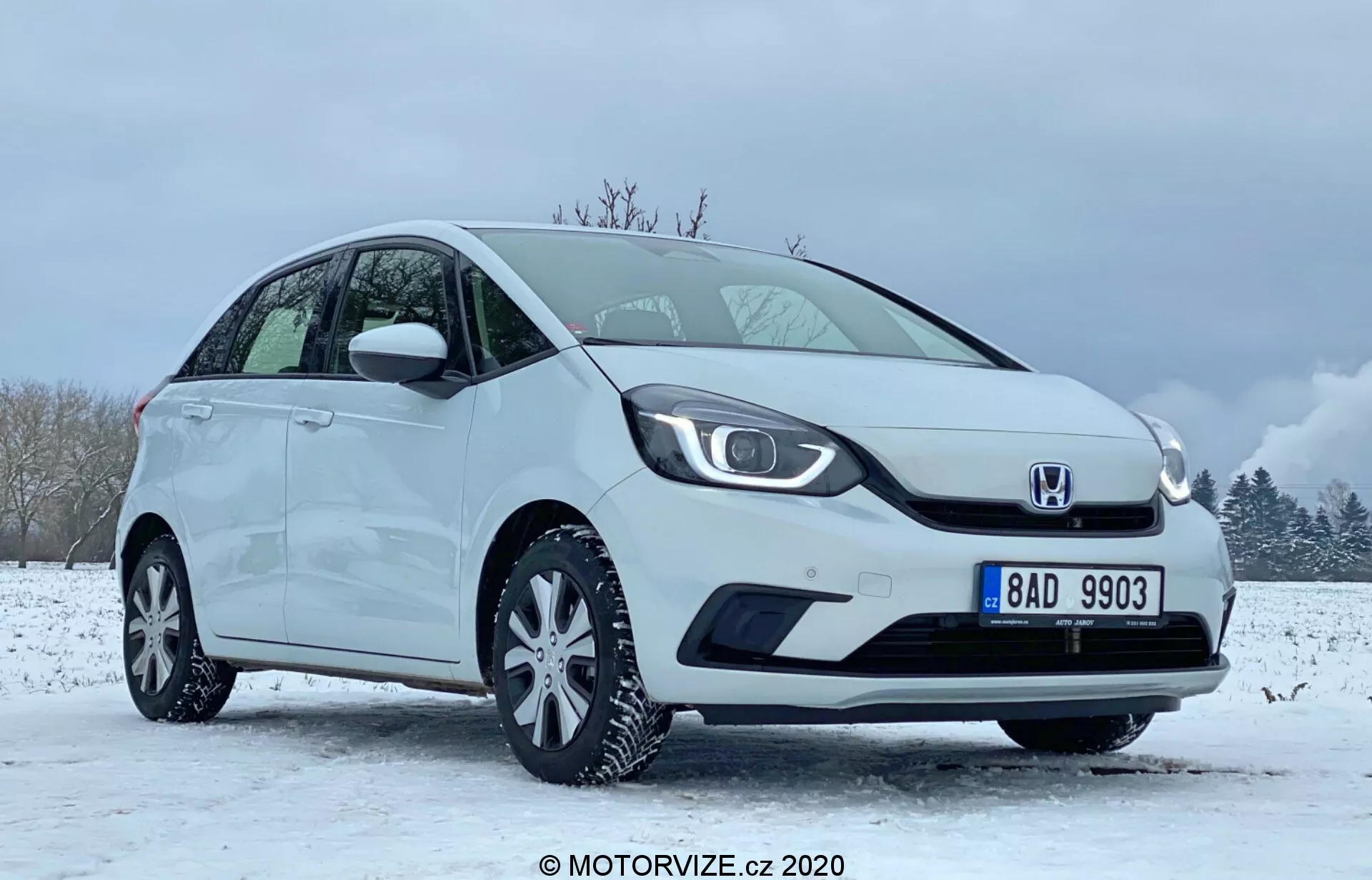 La imagen muestra un hatchback compacto Honda Jazz 2020 blanco posicionado en un entorno invernal con suelo cubierto de nieve y cielos parcialmente nublados. El coche se ve desde un ángulo de cuarto delantero centrado en el lado del conductor, destacando la parrilla distintiva con el logotipo en el centro, faros LED elegantes y parachoques delantero liso con carcasa de luces antiniebla integrada. El perfil lateral exhibe una línea de techo inclinada, llantas de aleación de múltiples radios, y espejos laterales y tiradores de las puertas del mismo color de la carrocería. El estado impecable del vehículo sugiere que podría ser una imagen promocional o de revisión capturada en 2020 por MOTORVIZE.cz.