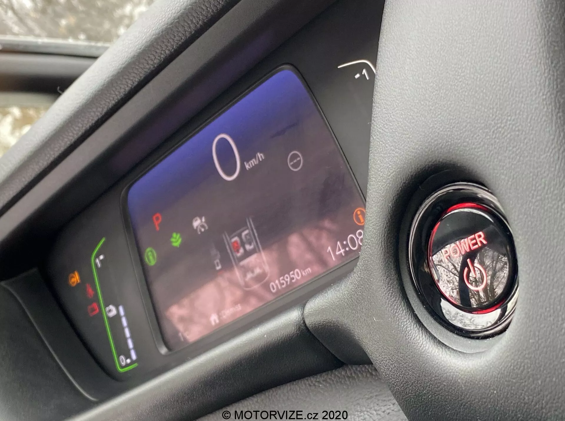 Vista de cerca del avanzado cuadro de instrumentos digital del conductor y el botón de arranque/parada del motor dentro del moderno interior del Honda Jazz 2020, tomada desde la perspectiva del asiento del conductor y enfocándose en los elementos del salpicadero. La pantalla digital del cuadro muestra un indicador de potencia con íconos ecológicos, un velocímetro central en 0 km/h, varias luces de advertencia, un reloj, y un odómetro con 015902 km. Junto a él se encuentra el botón de encendido del motor con un aro rojo para encendido sin llave.