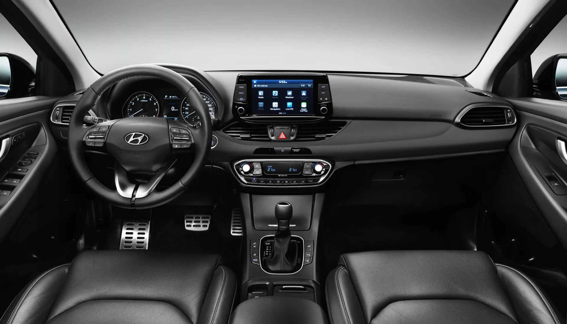 Interior del Hyundai i30 2017 Fastback mostrando el tablero frontal desde una perspectiva del asiento trasero, con el volante posicionado en el centro izquierda con botones de control y el logo de Hyundai, un cuadro de instrumentos digital detrás, un sistema de infotenimiento con pantalla táctil grande con aplicaciones de conectividad, control de clima de zona dual, palanca de transmisión automática, posibles controles de calefacción/enfriamiento de asientos y asientos tapizados en cuero.