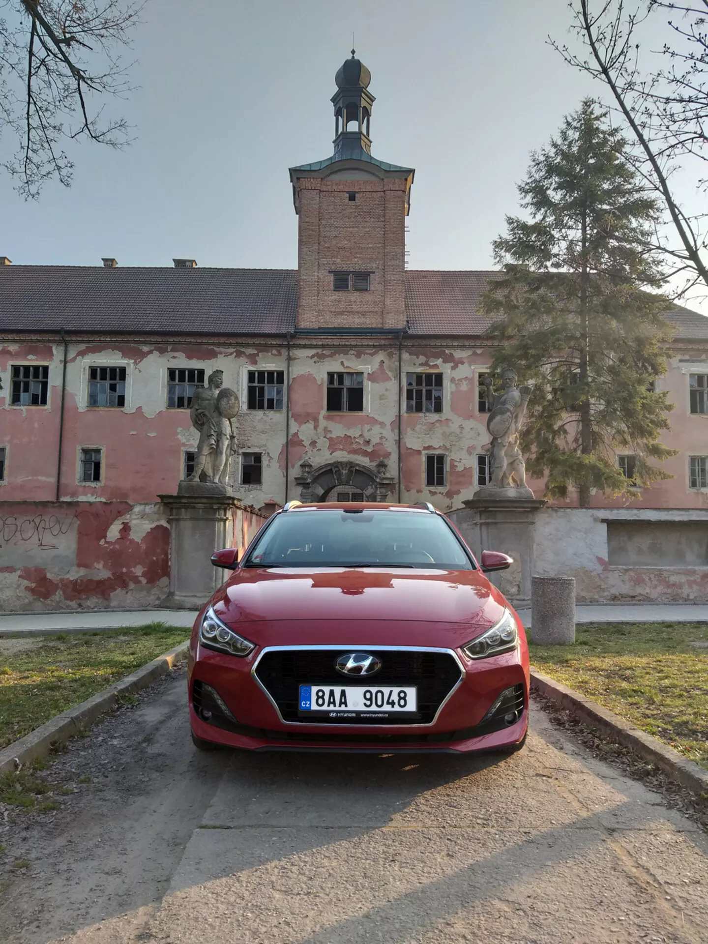 Hyundai i30 Fastback de 2017 en rojo de frente con vista completa de faros LED y diseño de parrilla esculpida, posicionado en un camino de grava con un edificio histórico de fondo.