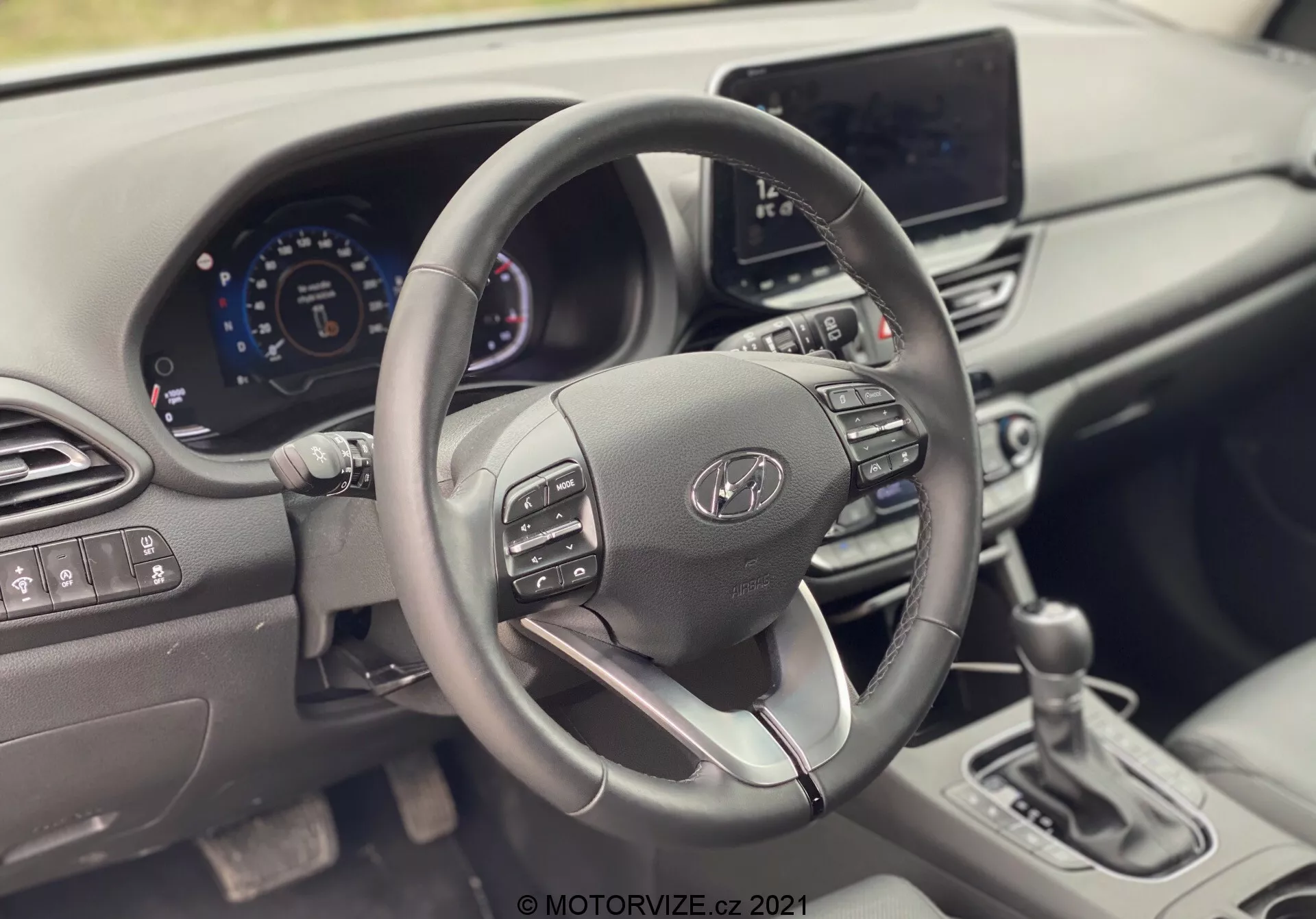 Vista interior del Hyundai i30 Kombi 2020 enfocada en el volante y el tablero. La fotografía está tomada desde el asiento del pasajero, mostrando un volante elegante con botones de control, un cuadro de instrumentos digital y un sistema de infoentretenimiento. El diseño moderno incluye un volante forrado en cuero con un radio inferior plateado, ventilaciones y controles físicos mínimos, destacando tecnologías automotrices avanzadas y diseño ergonómico.