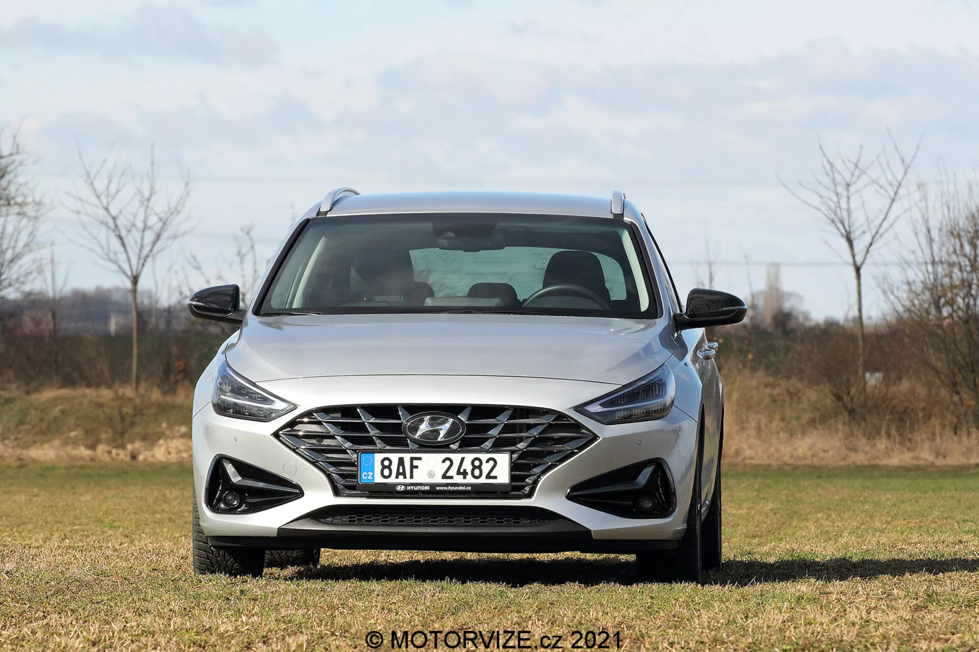 Vista en ángulo frontal de un Hyundai i30 2020 Kombi, con parrilla de patrón poligonal con el logo del fabricante, faros angulares afilados, parachoques delantero esculpido, líneas sutiles en el capó y espejos laterales en un área de hierba y fondo natural.