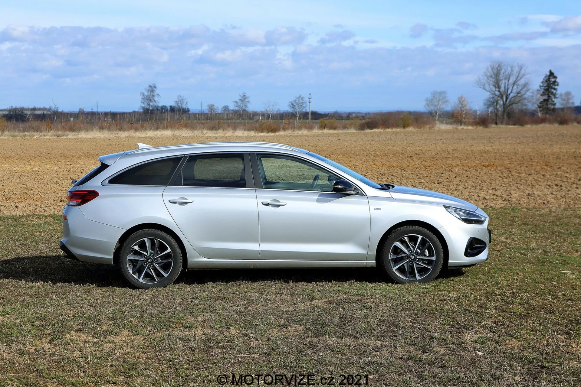 Perfil lateral de un Hyundai i30 familiar 2020 en color blanco brillante, estacionado en un campo de hierba seca con un paisaje árido y un cielo despejado de fondo. El vehículo cuenta con una parte trasera alargada para mayor espacio de carga, cuatro puertas, llantas de aleación de múltiples radios, grandes ventanas, espejos laterales, acabados cromados o metálicos, y rieles de techo integrados.