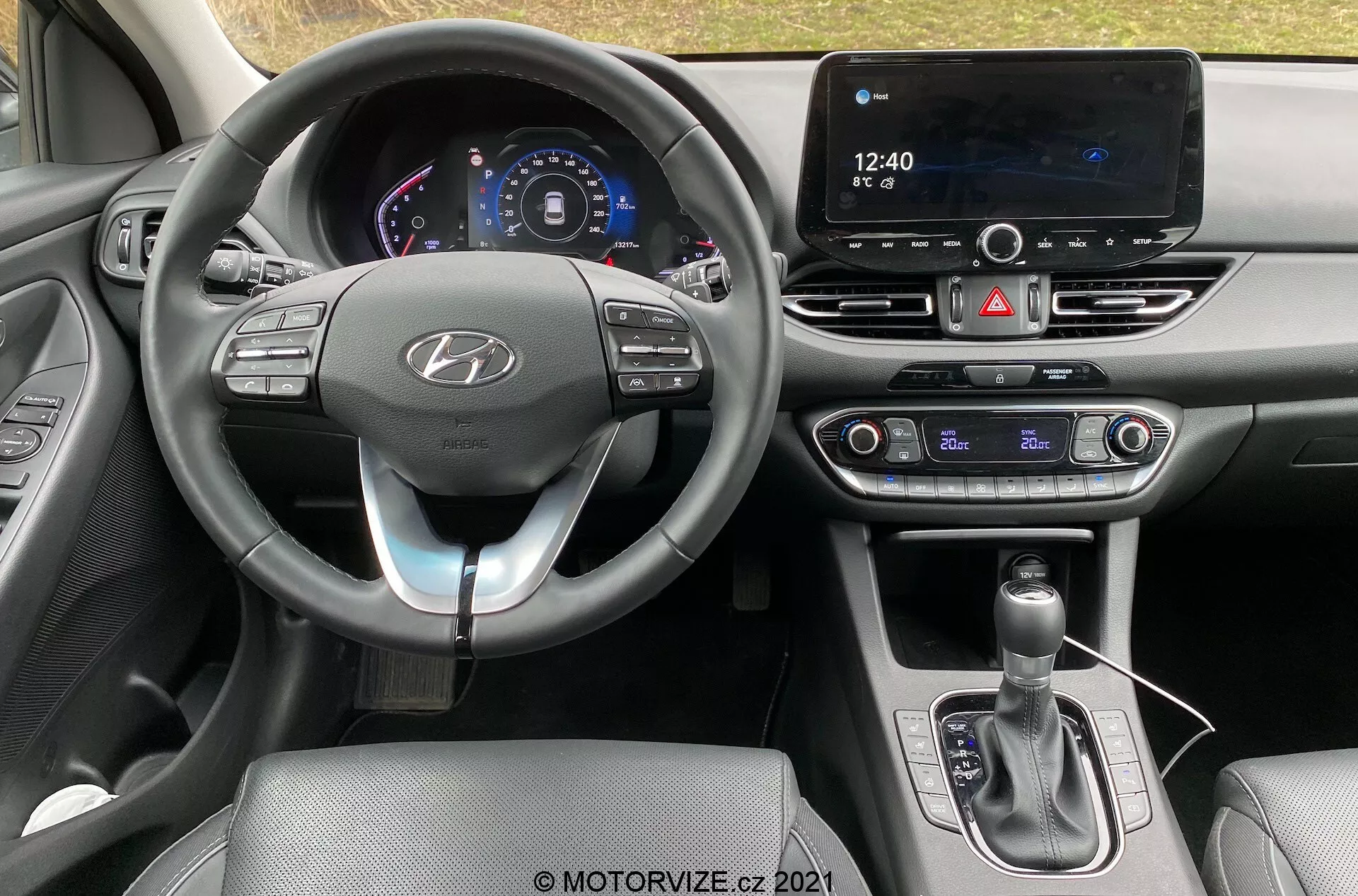 Vista del interior del Hyundai i30 Kombi 2020 desde el asiento del conductor, mostrando un moderno salpicadero con un volante adornado con el logo de Hyundai que cuenta con botones de control para funciones como control de crucero, audio y llamadas manos libres. Un cuadro de instrumentos digital muestra la velocidad y otra información del vehículo. El salpicadero central destaca una gran pantalla táctil de infoentretenimiento para navegación y medios, junto a controles físicos para el aire acondicionado, sugiriendo un sistema de climatización de doble zona. La consola central incluye una palanca de cambios con funda de cuero y un puerto USB, enfatizando características avanzadas de conectividad y tecnología.