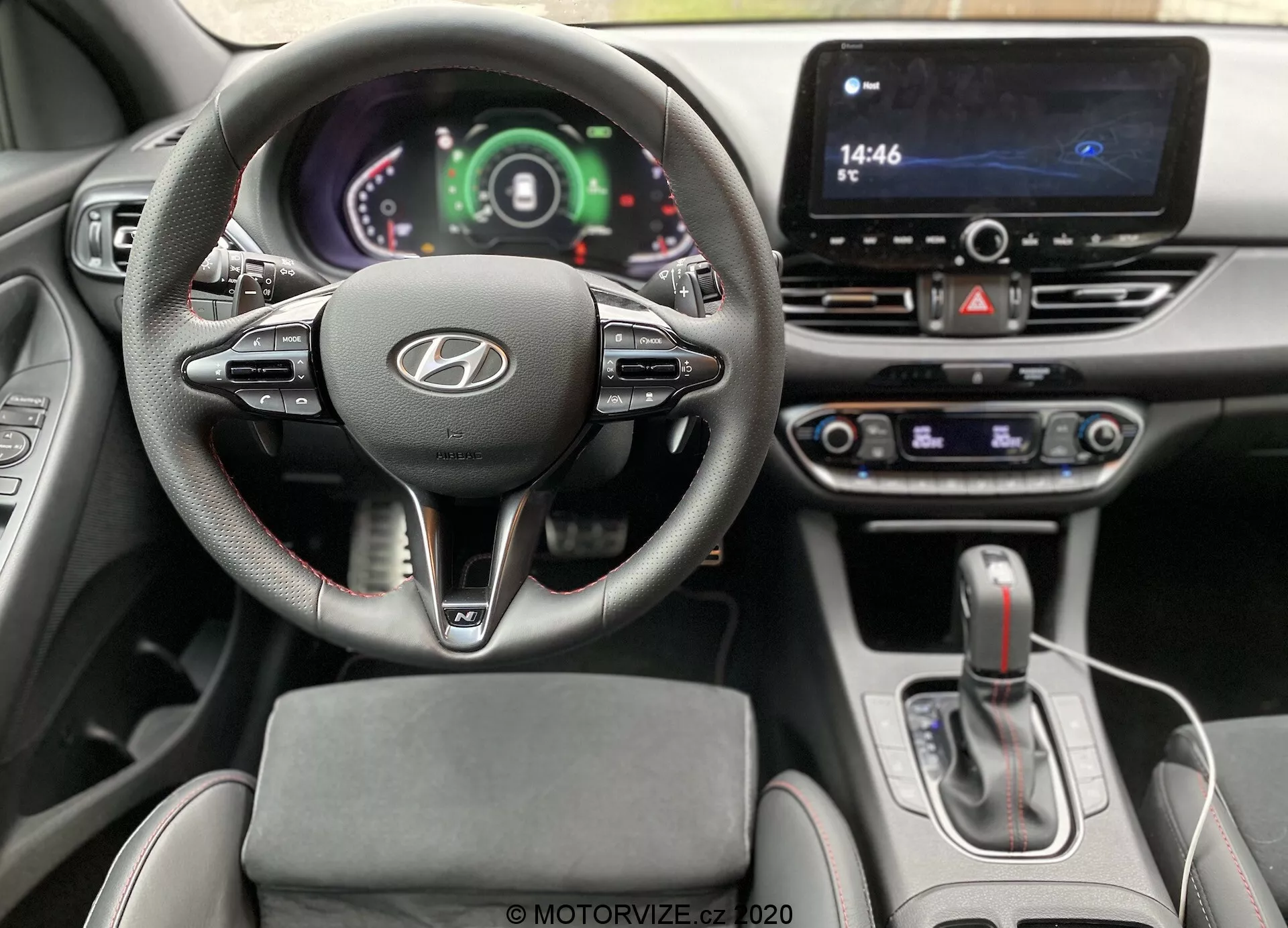 Vista interior desde la perspectiva del conductor dentro de un Hyundai i30 2020 (i30-kombi) que muestra el volante con el logo de Hyundai, controles montados y panel de instrumentos digital. Se pueden ver el sistema de infoentretenimiento con pantalla táctil, el selector de cambios de transmisión automática y los mandos de control de clima, indicando tecnología avanzada y diseño moderno.