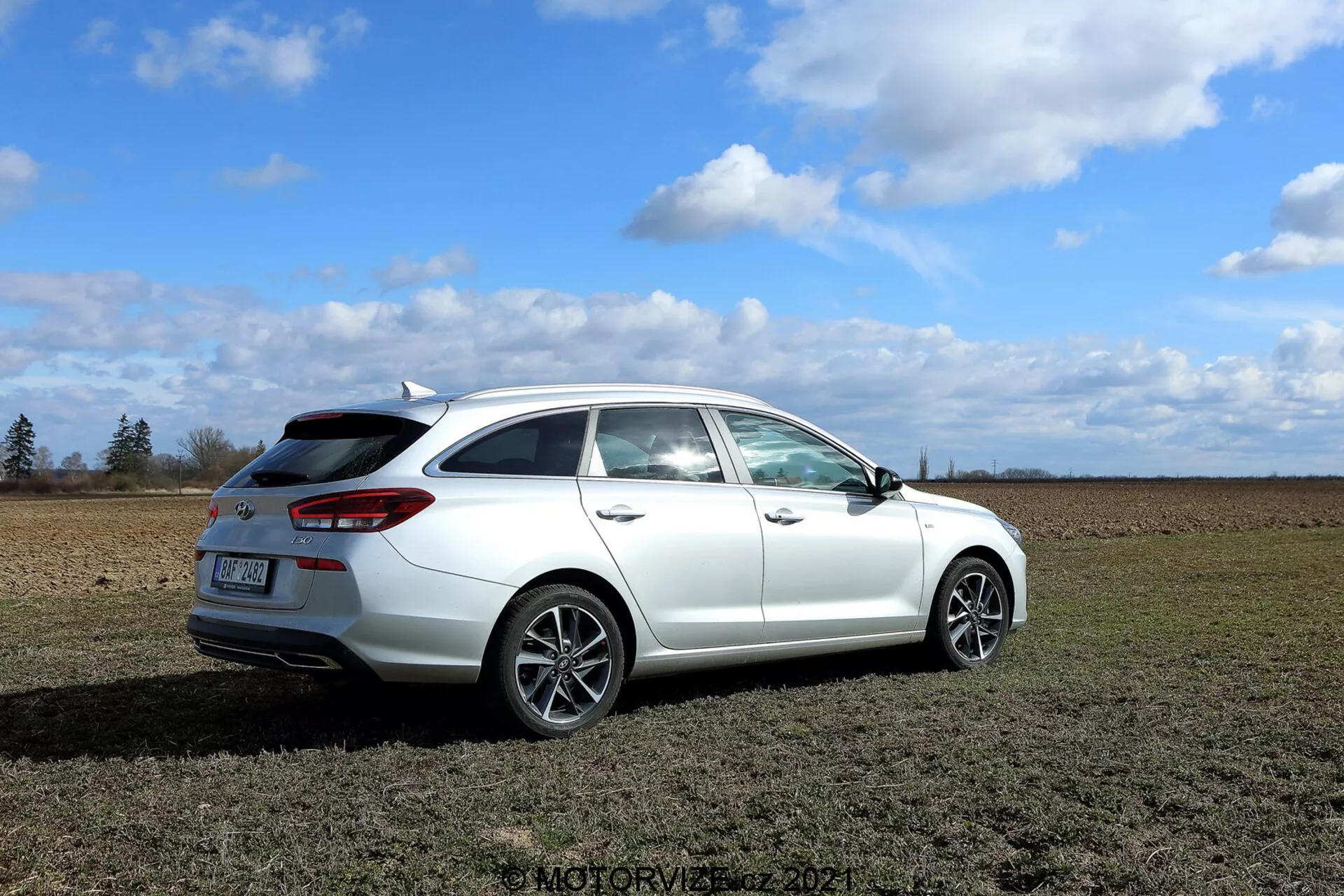 Una vista detallada de un Hyundai i30 station wagon blanco del 2020 estacionado en un campo de hierba con un cielo parcialmente nublado de fondo. La imagen está tomada desde un ángulo trasero de tres cuartos, mostrando elementos de diseño como las luces traseras modernas que envuelven los laterales del vehículo, el parabrisas trasero con limpiaparabrisas, el pequeño spoiler en el techo, las llantas de aleación multirradio y el espejo lateral con posibles características modernas. Las líneas de cierre de la puerta trasera de pasajeros y la puerta del maletero son visibles, indicando al menos cuatro puertas. Aunque el escape no es prominente, parece discreto. El coche está enfocado con nitidez, destacando su diseño y características, con el entorno proporcionando el escenario contextual.