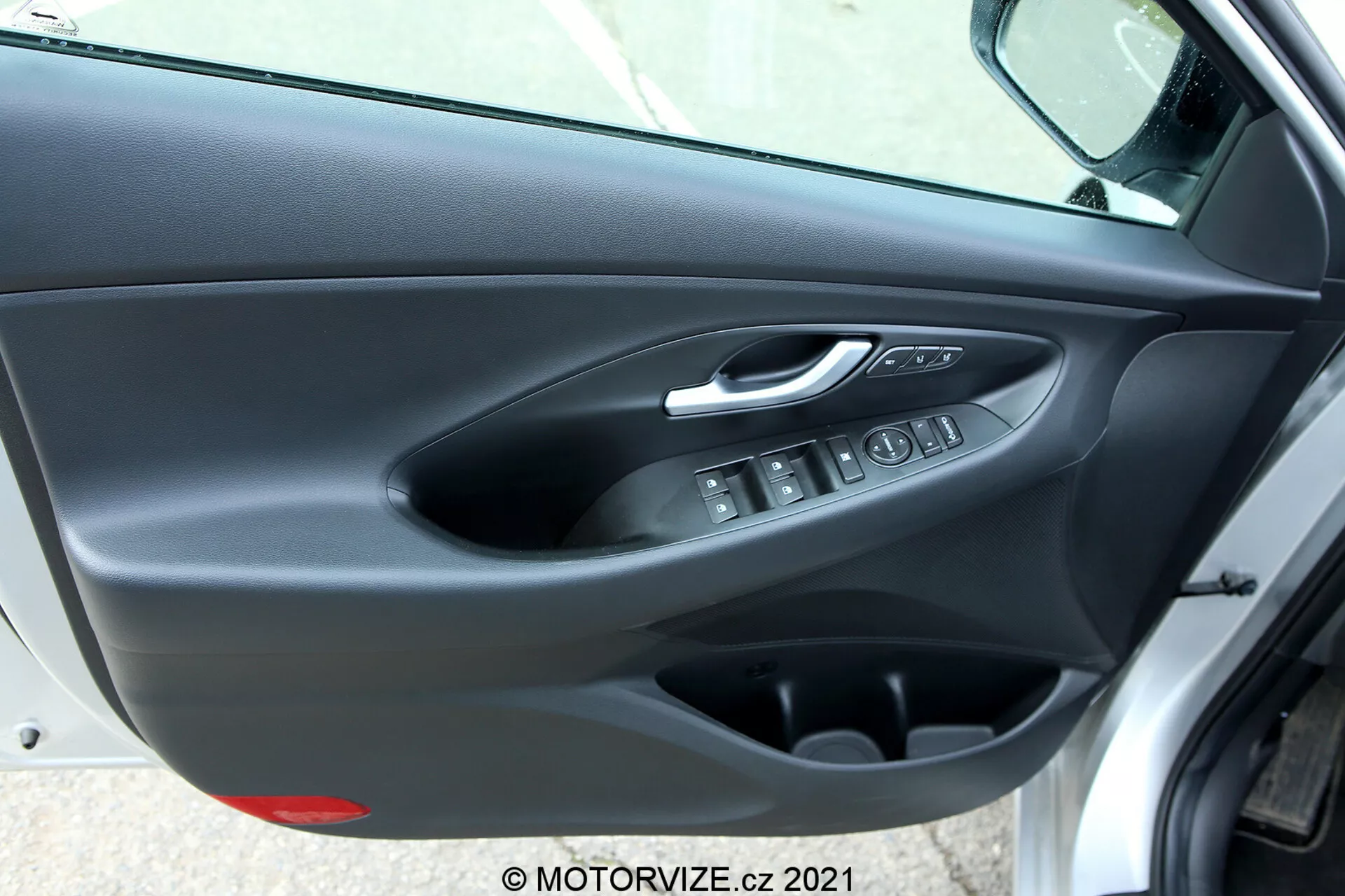 Panel de puerta interior del Hyundai i30 2020 (i30-kombi) visto desde un ángulo oblicuo, mostrando la manija de la puerta con acabado cromado plateado, botones de control ergonómicos para las ventanas eléctricas, botón electrónico de bloqueo de la puerta, bolsillo de almacenamiento y revestimiento de la puerta de doble textura con panel superior suave y parte inferior de plástico rígido en colores oscuros.