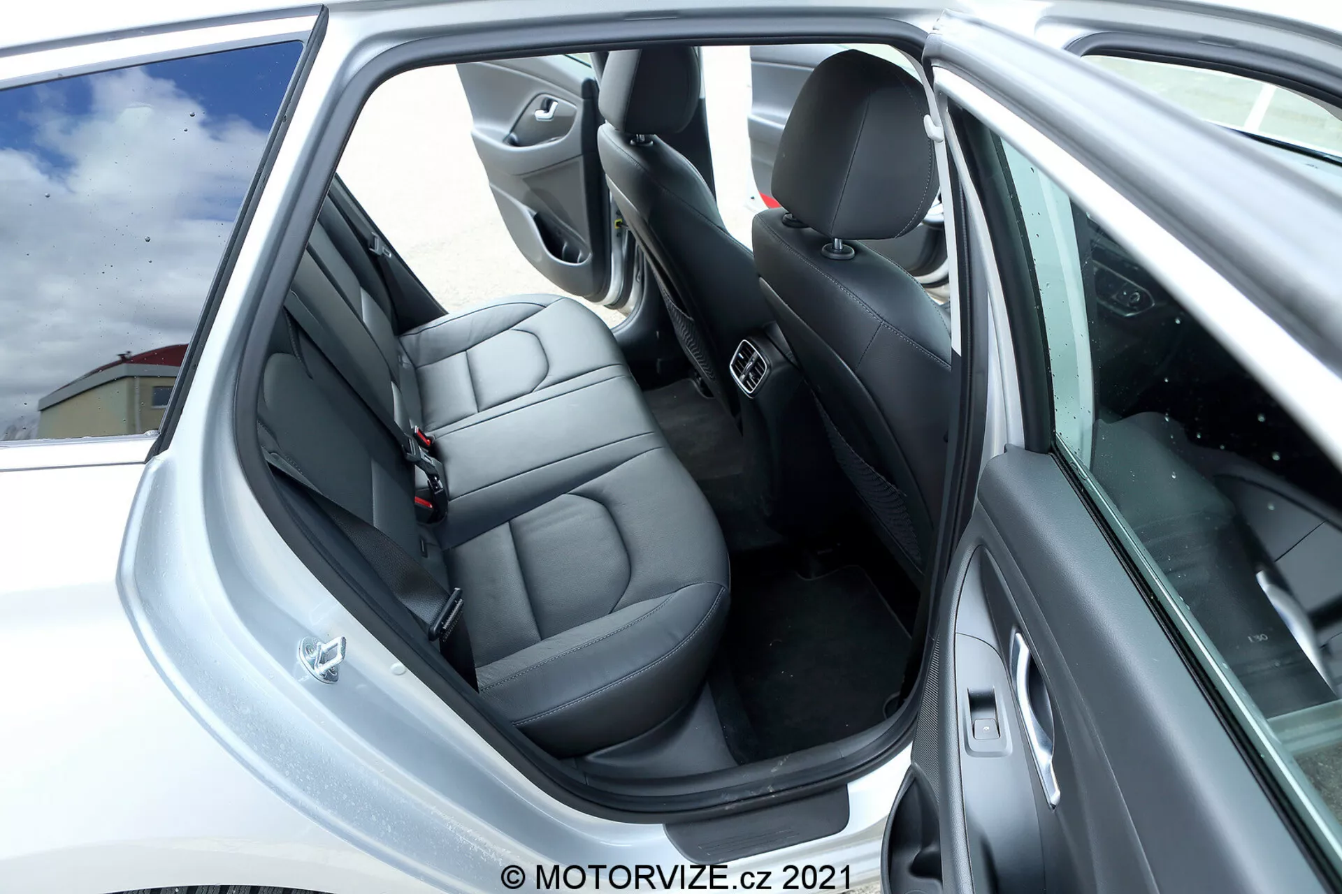 Vista de cerca del área de los pasajeros traseros del Hyundai i30 2020. La imagen muestra la puerta trasera abierta con una vista clara de los asientos traseros de cuero oscuro, que tienen reposacabezas individuales y cinturones de seguridad. El panel de la puerta cuenta con acentos grises o plateados con botones de control y una rejilla de altavoz, indicando características tecnológicas y de audio. Los asientos delanteros y el pilar B son visibles, reflejando un diseño y material similar al de los asientos traseros. El interior, predominantemente negro con algunos acentos plateados, está bien mantenido, enfocado en la comodidad y el diseño.