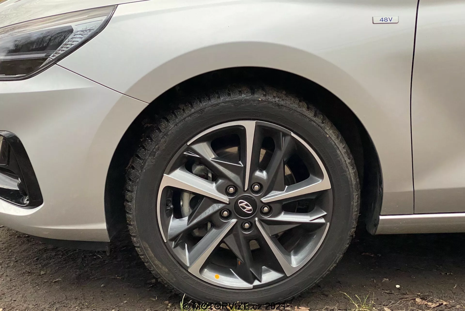 Primer plano de la rueda delantera del Hyundai i30 Kombi 2020, destacando su llanta de aleación moderna con un diseño de múltiples radios y frenos de disco visibles a través de los radios. La vista, tomada desde un ángulo bajo cerca del suelo, incluye parte del lateral del vehículo en color plata metálico, con una insignia 4WD que indica la capacidad de tracción a las cuatro ruedas. La imagen resalta el diseño limpio del coche con líneas curvas y detalles característicos menores sobre el arco de la rueda.