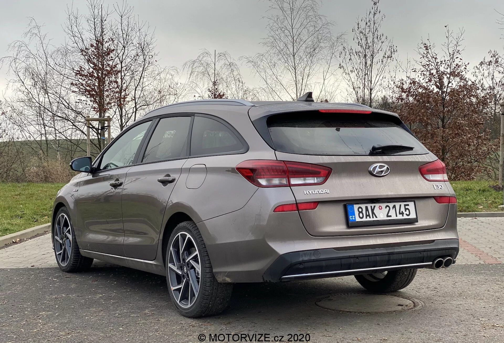 Imagen de un Hyundai i30 2020 gris (i30-kombi) desde una perspectiva trasera de tres cuartos. El coche está estacionado al aire libre en un entorno suburbano o de parque, mostrando las características de diseño traseras y laterales del pasajero. Elementos visibles incluyen un maletero con alerón, luces traseras envolventes, insignia 'i30', insignia 'N Line', tubos de escape dobles y llantas de aleación de varios radios. El entorno sugiere un área con césped y árboles en el fondo.
