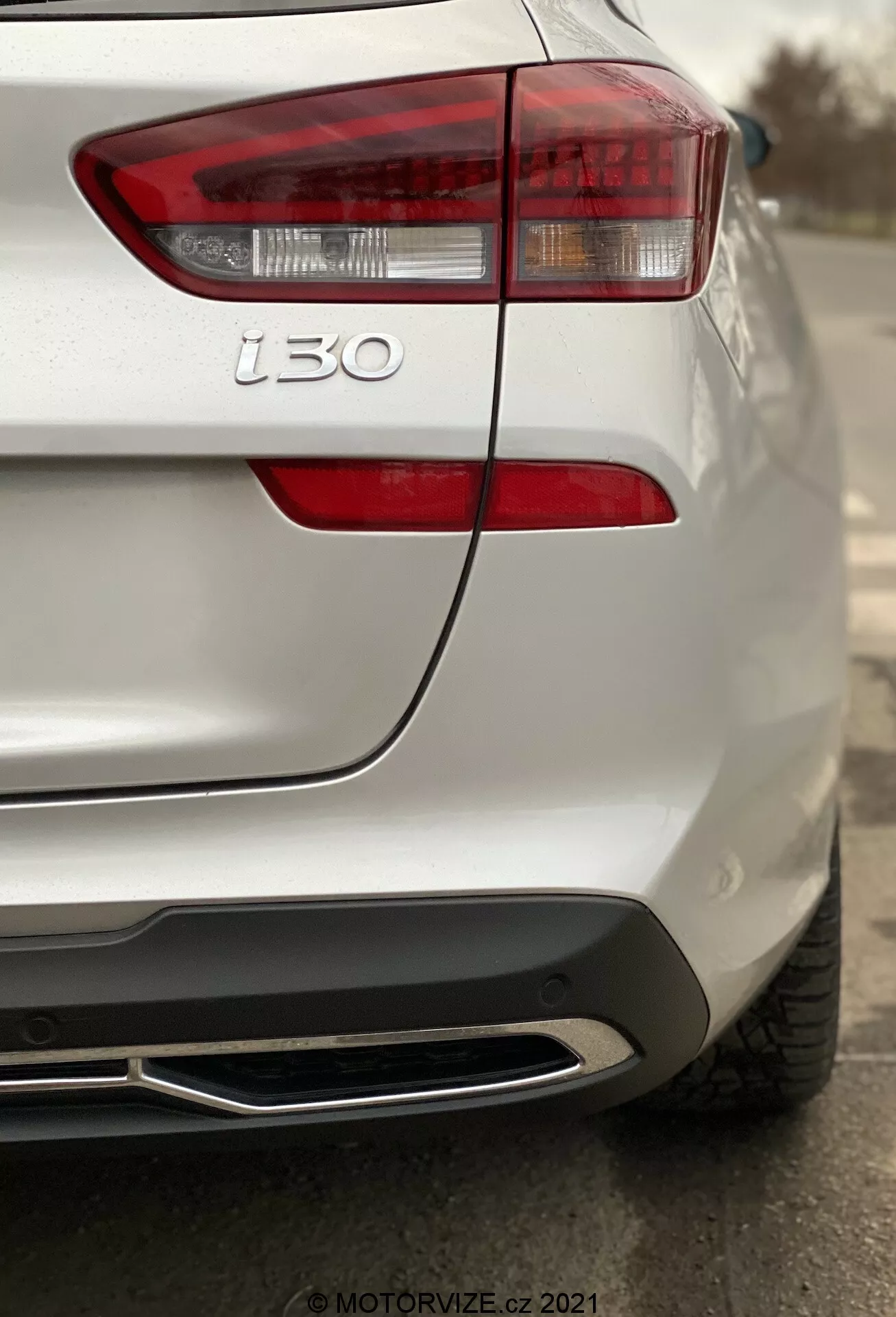 Vista trasera en primer plano de un Hyundai i30 2020 (Kombi) blanco enfocada en el conjunto de la luz trasera izquierda y sección trasera izquierda, mostrando líneas de diseño moderno, luces de freno con lente claro, intermitentes, insignia i30, textura detallada de la pintura, sensor de aparcamiento, integración de diseño a ras y salida de escape con punta de cromo, con poca profundidad de campo resaltando el diseño trasero del coche.