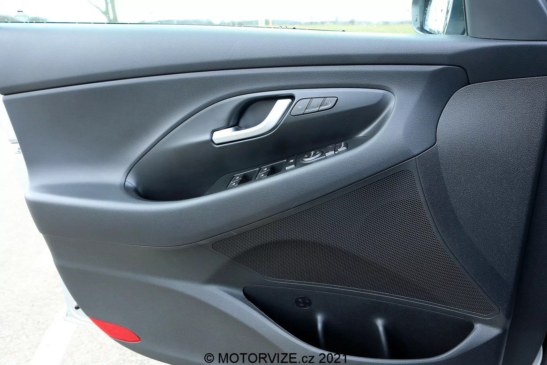 Primer plano del panel interior de la puerta del lado del conductor del Hyundai i30 Kombi 2020 que muestra la manija de la puerta plateada, los controles de las ventanas y los espejos, el botón de bloqueo de las puertas, el altavoz integrado en la parte inferior y un acabado negro mate con textura con un pequeño compartimento de almacenamiento probablemente en la parte inferior.