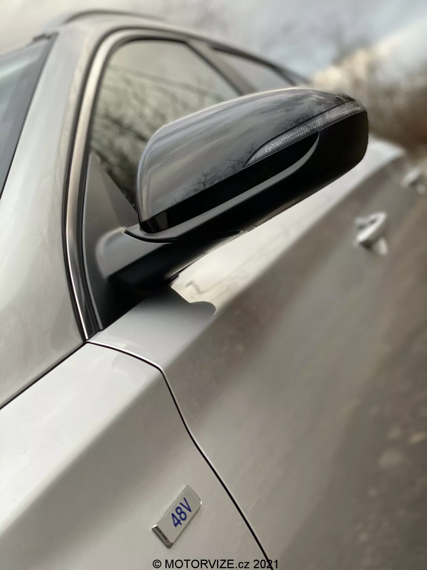 Imagen de primer plano en ángulo del espejo lateral del Hyundai i30 2020 tomada desde la parte trasera hacia el frente. La toma se enfoca en el conjunto del espejo, incluyendo su moderno alojamiento con indicadores de giro integrados y superficie reflectante. La puerta del conductor y el color exterior claro del coche están desenfocados, lo que resalta el diseño y los detalles del espejo. La insignia '4DV' es visible pero su significado específico no está claro.