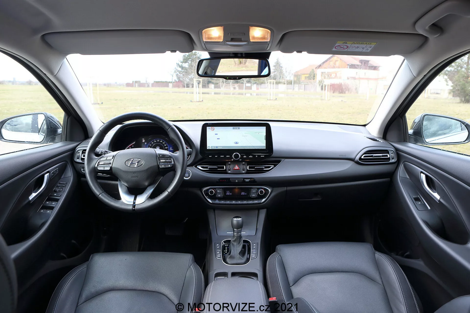 Interior del Hyundai i30 2020 Kombi que muestra el área del tablero delantero, tomada desde los asientos traseros. La imagen captura un volante multifunción moderno con botones de control, un sistema de infoentretenimiento con pantalla táctil que incluye navegación y opciones multimedia, control de clima de doble zona, una palanca de cambios automática y asientos delanteros ergonómicos con reposacabezas. El diseño interior de dos tonos contrasta tonos oscuros y claros, añadiendo profundidad. Los paneles de las puertas reflejan el diseño con posibles rejillas de altavoces y botones de control para ventanas y espejos eléctricos. La consola superior incluye luces de cabina y botones para funciones adicionales.