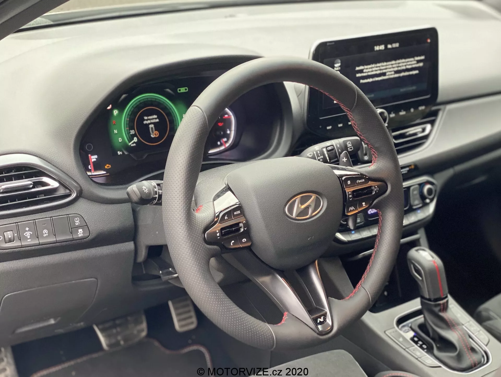 Vista interior de un Hyundai i30 2020 (i30-kombi) desde el asiento del conductor, enfocándose en el volante y el tablero. El volante moderno cuenta con múltiples botones de control para volumen, comandos de voz y llamadas manos libres. El panel de instrumentos combina elementos digitales y analógicos, indicando sistemas avanzados de asistencia al conductor. Una pantalla táctil de infoentretenimiento central es visible, lo que sugiere navegación y reproducción de medios. La consola central muestra botones de control del clima y una palanca de cambios para transmisión automática. Controles de ventanas eléctricas y espejos laterales están en la puerta del conductor.