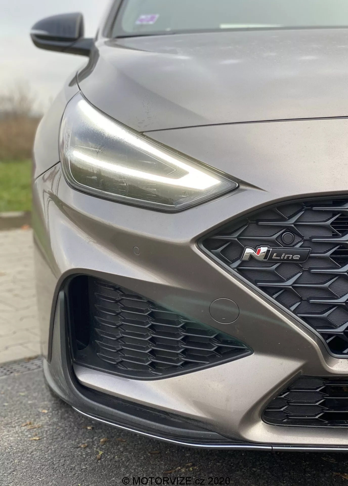 Vista cercana y angular enfocada en el lado delantero izquierdo de un Hyundai i30 2020 N Line gris. La imagen resalta el diseño deportivo, incluyendo un sutil contorno del capó, faros LED afilados, una parrilla en forma de panal con una prominente insignia roja 'N Line' y un intrincado parachoques con sensores avanzados que sugieren sistemas modernos de asistencia a la conducción.