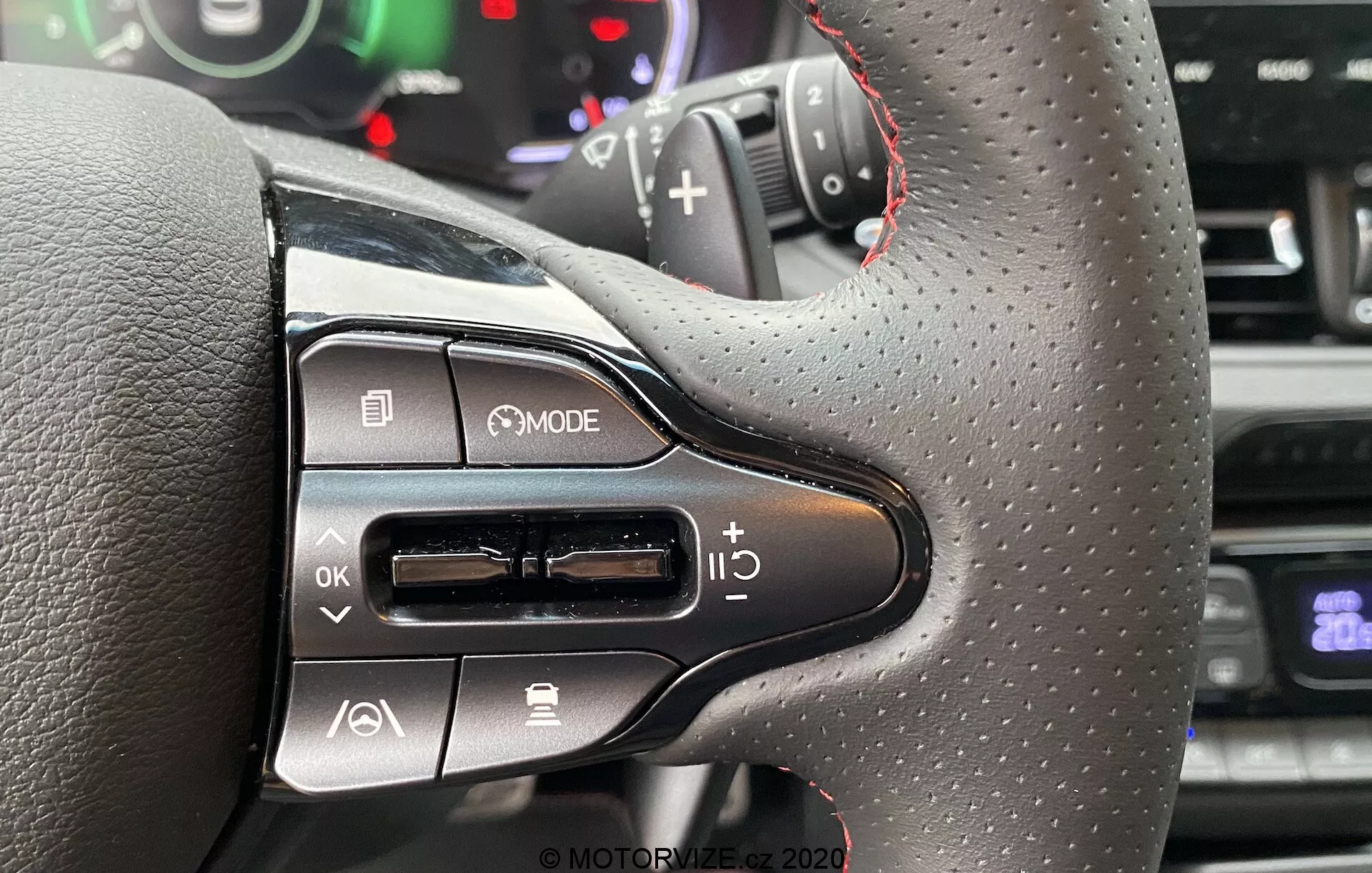 Primer plano de la parte izquierda del volante del Hyundai i30 Kombi 2020 con textura similar a cuero y costuras rojas, controles multifuncionales modernos incluido un botón MODE, un pad de navegación, control de volumen y vista parcial de la paleta de cambios. El fondo muestra panel de instrumentos o indicadores borrosos indicando la perspectiva del conductor.
