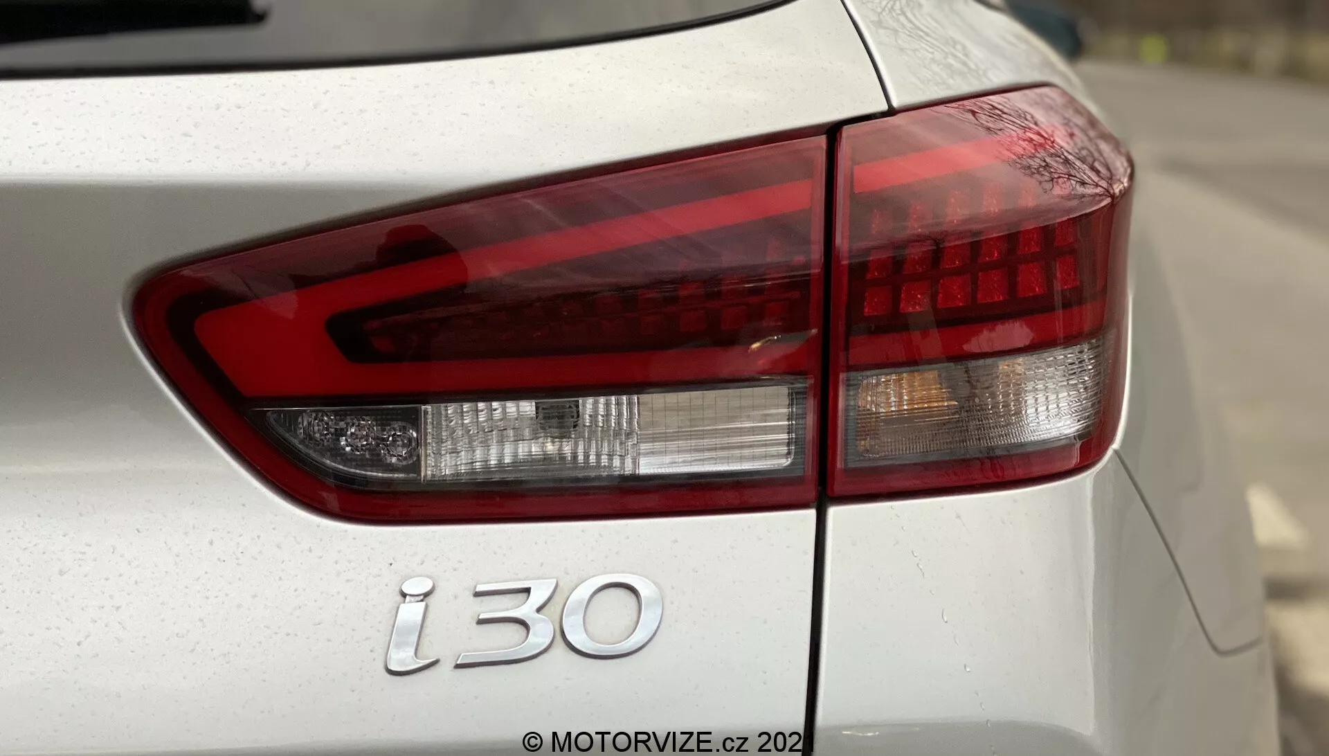 Primer plano del lado trasero izquierdo de un Hyundai i30 Kombi 2020, destacando el conjunto de luces traseras modernas con luz de freno roja, luz de reversa blanca e indicador ámbar bajo especificaciones europeas. La toma incluye parte de la tapa del maletero, el parachoques trasero y el emblema 'i30', con pintura probablemente de color plateado o blanco. La poca profundidad de campo enfatiza las luces traseras y el emblema, con un fondo borroso y la marca de agua 'MOTORVIZE.cz 202' visible.