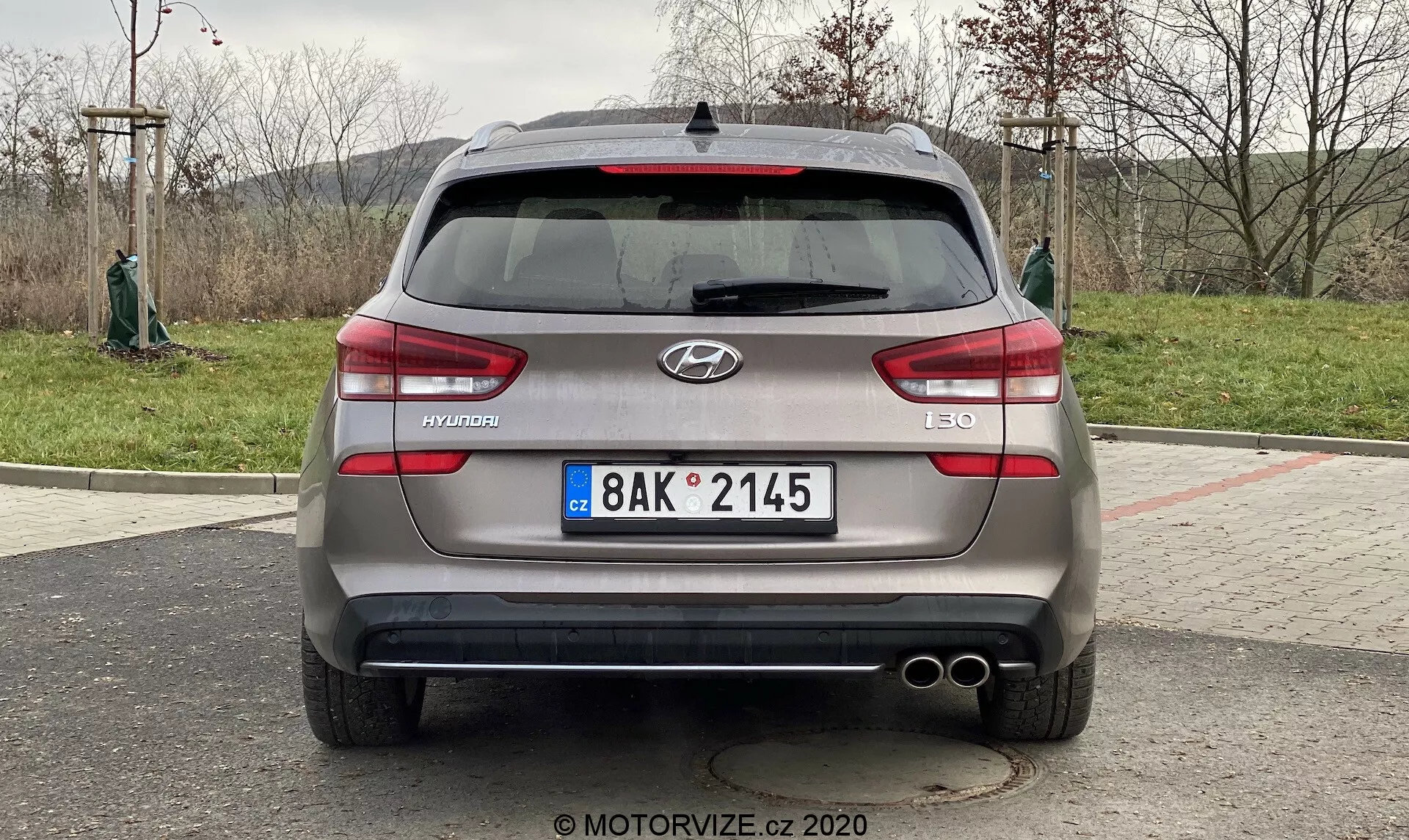 La imagen muestra la vista trasera de un Hyundai i30 Kombi 2020. El coche está aparcado y la toma se realiza desde un ángulo directo, aproximadamente a la altura del maletero del vehículo. Centrada en el vehículo, la imagen proporciona una vista clara y simétrica de las características traseras del coche, incluyendo el logo de Hyundai, el nombre del modelo 'i30' y el nivel de acabado 'CRDi' que indica un motor diésel Common Rail de inyección directa. Los detalles visibles incluyen la matrícula de estilo europeo, luces traseras horizontales, sensores traseros, doble salida de escape, limpiaparabrisas trasero, antena de aleta de tiburón y posiblemente una cámara trasera cerca del área de la matrícula.
