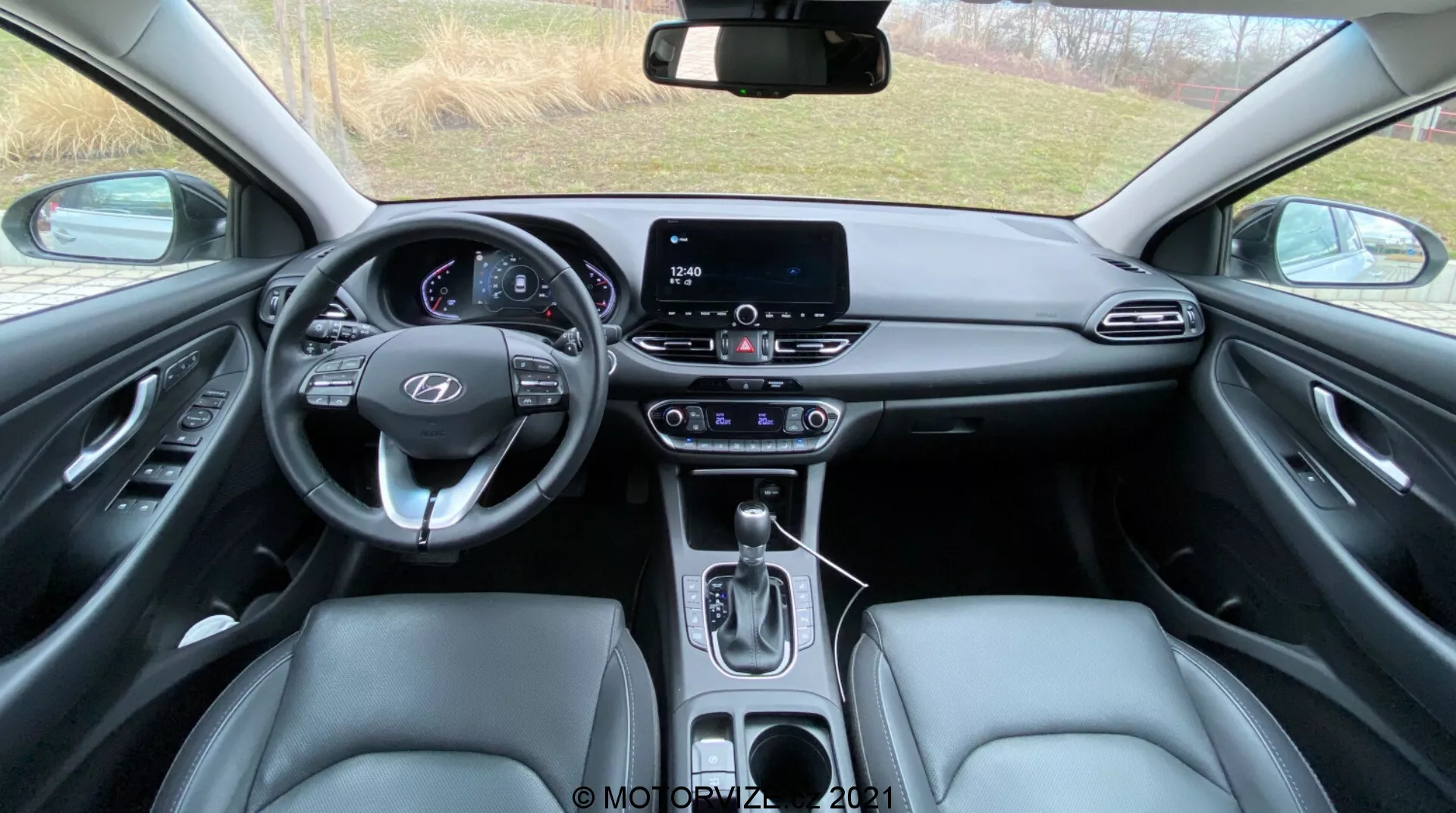 Vista interior de un Hyundai i30 2020 (i30-Kombi) desde el asiento trasero, mostrando el volante multifuncional con controles montados, panel de instrumentos digital que indica el modo de estacionamiento y la puerta del conductor abierta, sistema de infoentretenimiento de pantalla táctil, palanca de cambios automática, asientos delanteros tapizados en cuero, paneles de las puertas con controles eléctricos, materiales interiores diversos que incluyen cuero sintético y acentos metálicos, consola superior con espacio para gafas de sol y botones, espejo retrovisor autodimming con botones adicionales y alfombrillas de moqueta parcialmente visibles.
