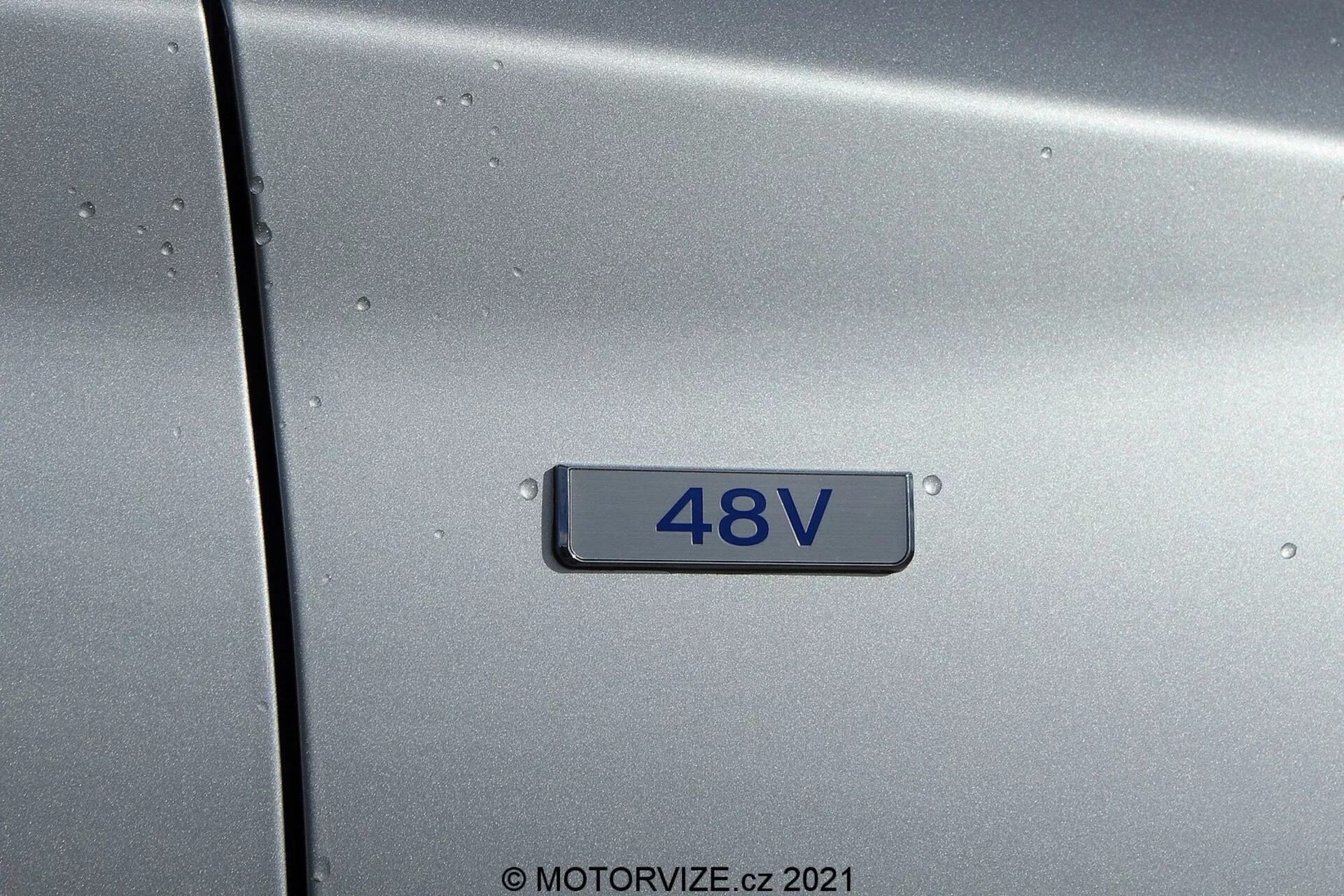 Vista de cerca del panel lateral de un Hyundai i30 2020 (i30-Kombi) color gris metálico que muestra una insignia '48V', indicando un sistema híbrido suave de 48 voltios, con superficie salpicada por gotas de lluvia, sin vista panorámica, iluminación neutra, fondo negro y números plateados.