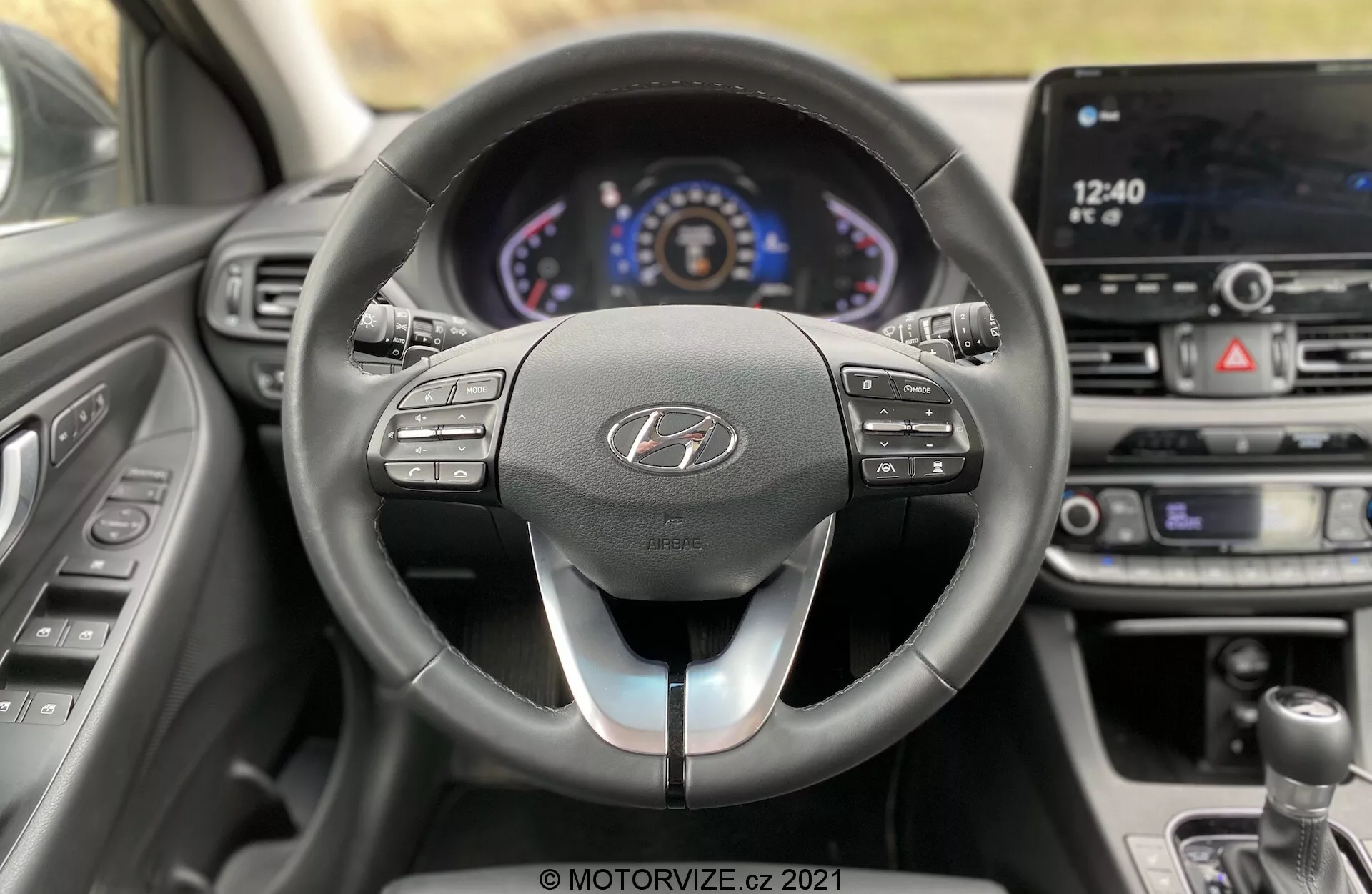 Vista interior del Hyundai i30 Kombi 2020 desde la perspectiva del conductor, mostrando un moderno habitáculo con un volante de tres radios, cuadro de instrumentos digital y pantalla táctil de infoentretenimiento. El volante cuenta con múltiples botones de control para audio, comunicación y control de crucero con un diseño bicolor. El cuadro de instrumentos iluminado muestra un velocímetro, tacómetro y pantalla de información central. El tablero central incluye ventilaciones y control de clima. La vista parcial de la palanca de cambios sugiere una transmisión manual. El coche parece estar detenido sin desenfoque de movimiento visible a través de las ventanas.