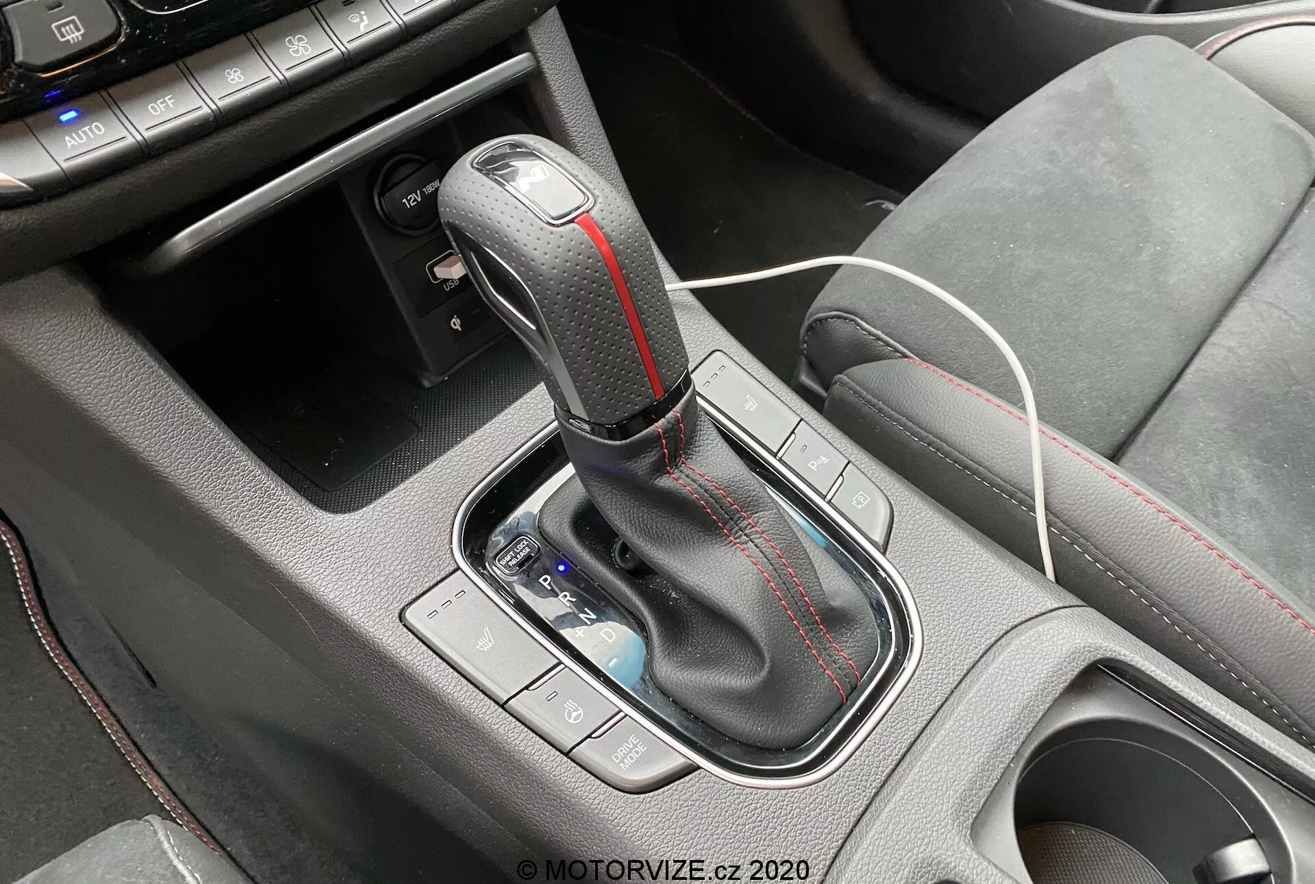 Primer plano del área de la consola central de un Hyundai i30 2020 kombi, con la palanca de cambios automática. La palanca tiene un pomo envuelto en cuero con acentos metálicos y costuras en contraste rojo. Un pequeño espacio de almacenamiento con un cable de carga blanco es visible delante de la palanca, indicando la presencia de puertos USB o una toma de corriente de 12V. El área circundante de la consola está hecha de acabados de plástico texturizado para un mejor agarre. Los controles a la izquierda de la palanca, probablemente para modos de conducción o control de tracción, están ligeramente desenfocados.