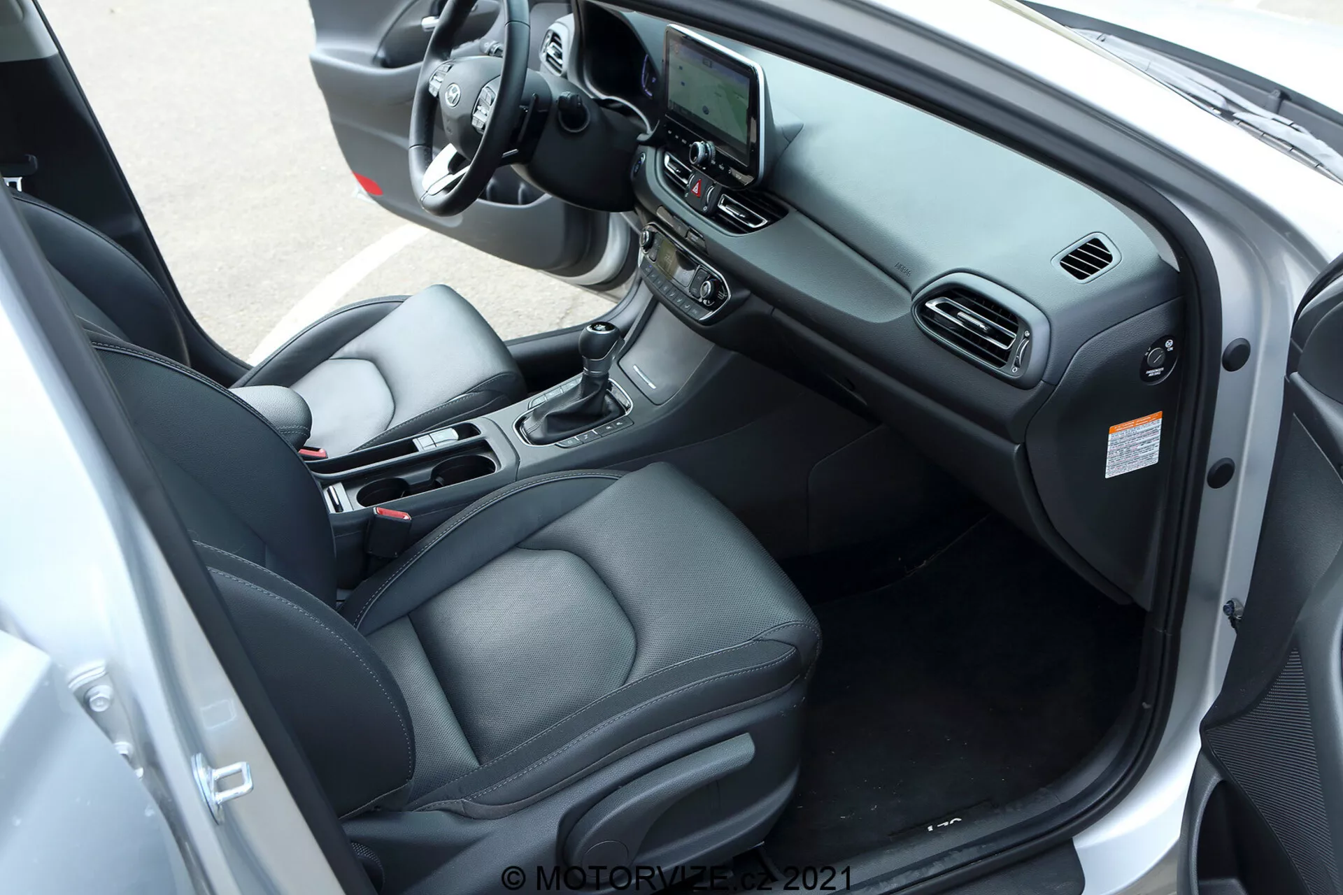 Interior del Hyundai i30 kombi 2020 capturado desde un ángulo diagonal y ligeramente elevado centrado en el lado del conductor. La imagen muestra el asiento del conductor tapizado en cuero con controles de ajuste electrónico, un volante multifuncional con varios botones de control, un sistema de infoentretenimiento central con pantalla táctil y una palanca de cambios automática en la consola central. Otros elementos incluyen portavasos, un compartimento de almacenamiento, control de clima automático y controles para ventanas eléctricas y espejos laterales en el panel de la puerta del conductor. La puerta del automóvil está abierta de par en par para proporcionar una vista completa del tablero, el diseño de los asientos y los elementos de tecnología y confort del interior.