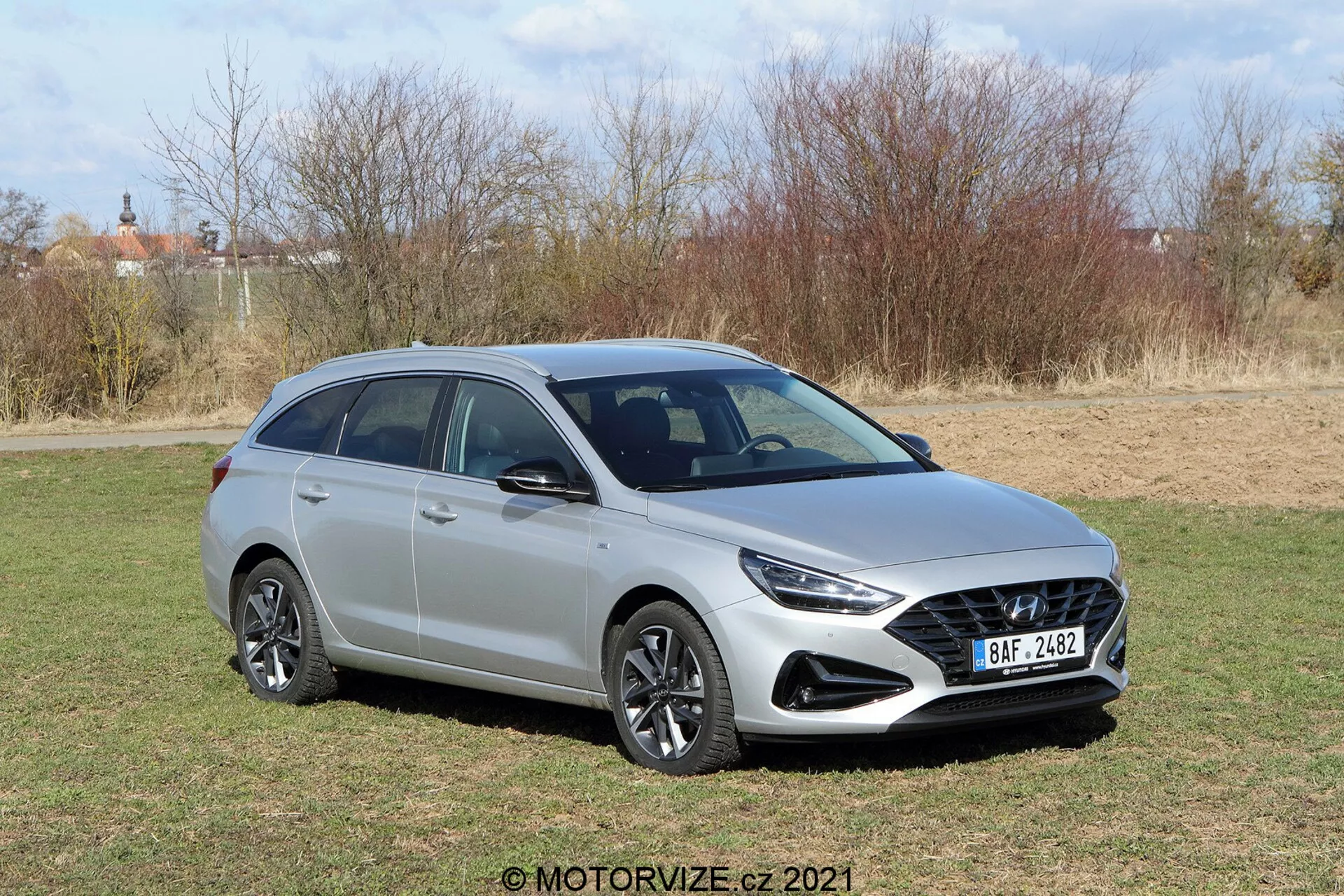 Vista de tres cuartos frontal de un moderno Hyundai i30 2020 station wagon hatchback, tomada desde un ángulo elevado que muestra la parrilla delantera con diseño de panal, faros elegantes, llantas de aleación de diseño multirradio, espejos laterales con señales de giro integradas, y rieles en el techo sobre una carrocería metálica con líneas suaves y esculpidas en un entorno natural soleado.