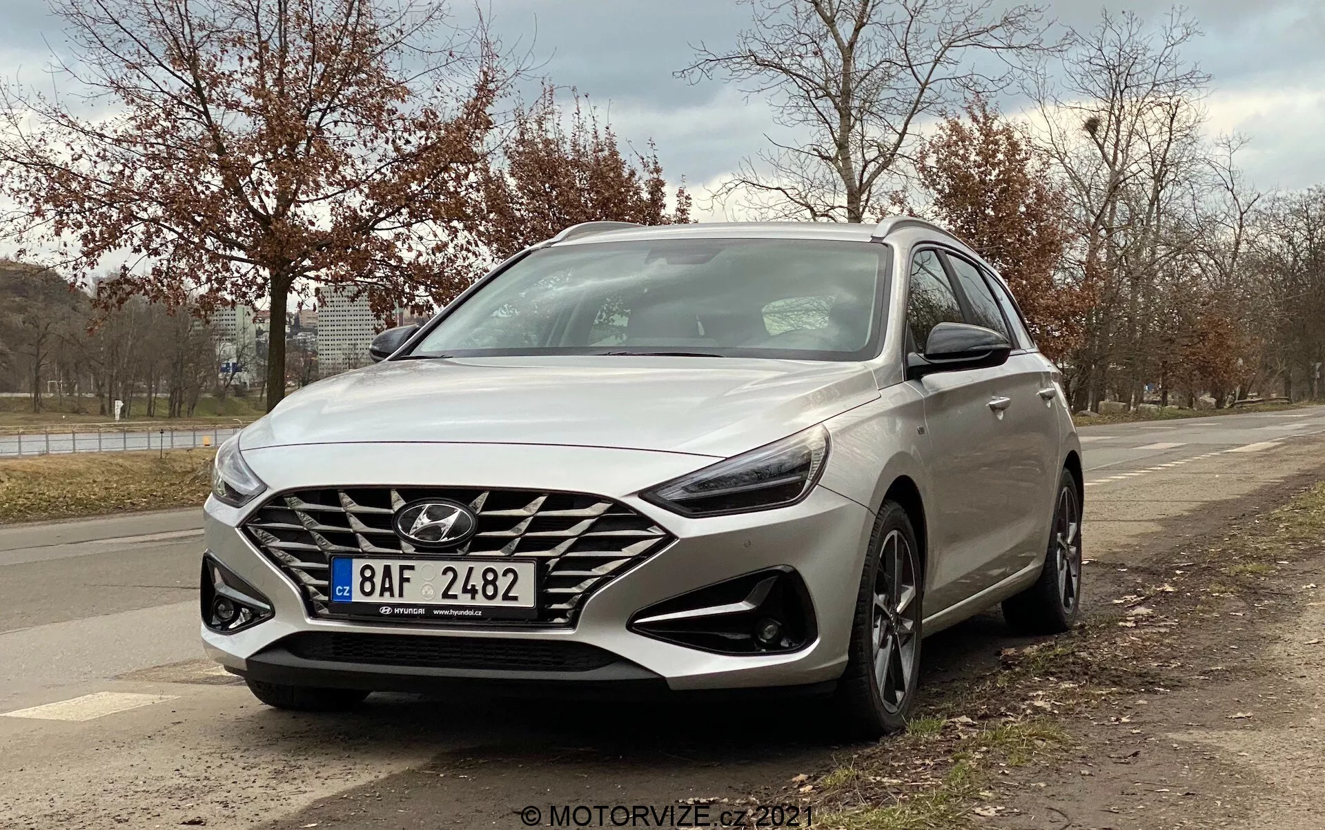 La imagen presenta un Hyundai i30 Kombi 2020 plateado capturado desde un ángulo bajo de tres cuartos desde el frente, mostrando el diseño desde el lado del conductor. El ángulo destaca la gran parrilla en cascada con el prominente logotipo de Hyundai, los faros LED modernos, las llantas de aleación de varios radios, las luces diurnas integradas debajo de los faros y un sensor de radar visible para las funciones de seguridad. También se destacan las líneas y contornos aerodinámicos del coche, con espejos laterales que incorporan señales de giro. El fondo incluye una acera, árboles y un cielo nublado, sugiriendo un entorno suburbano al aire libre.