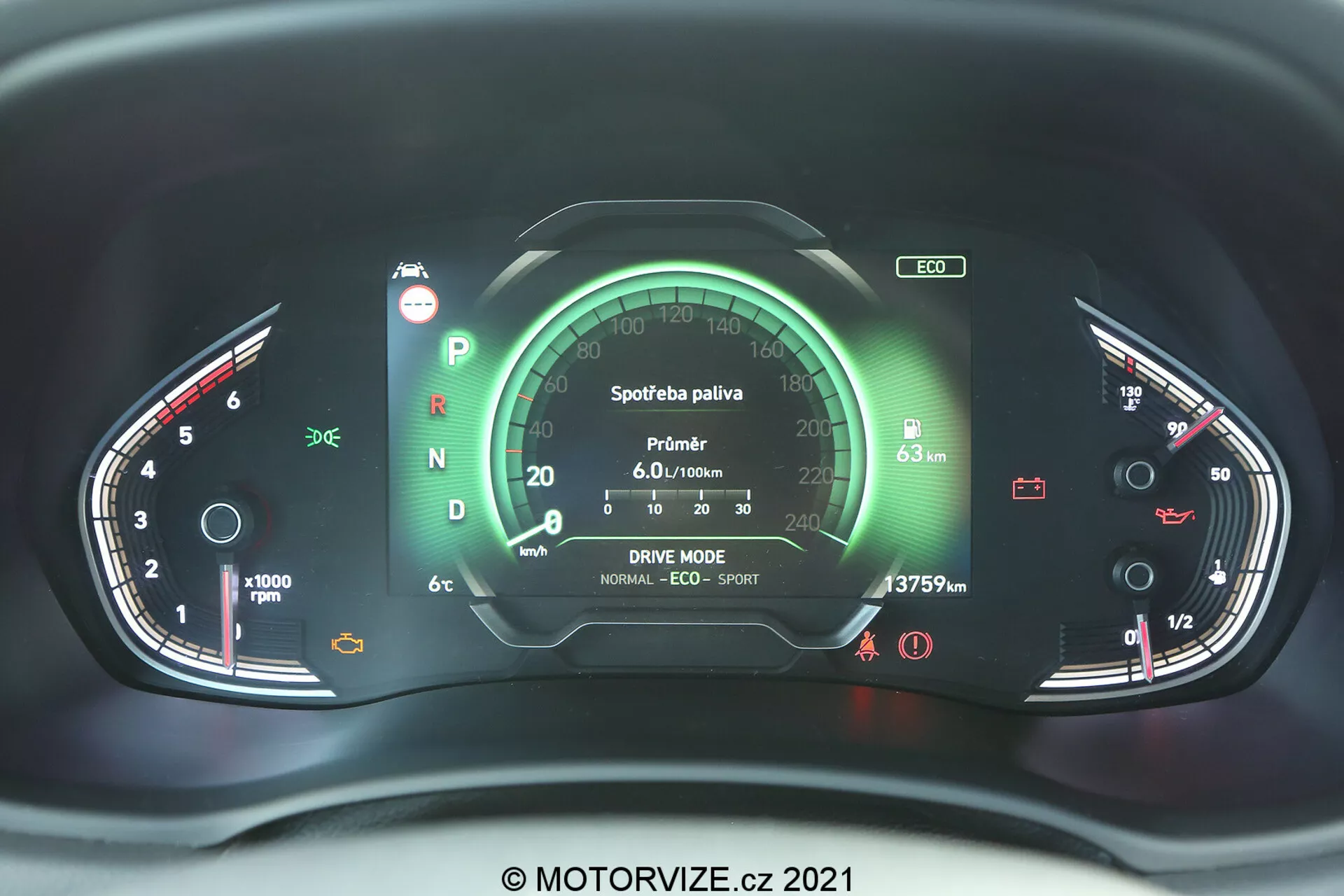 Vista de cerca del cuadro de instrumentos moderno del Hyundai i30 2020 desde la perspectiva del conductor, con una pantalla central digital flanqueada por medidores analógicos. El lado izquierdo muestra un tacómetro, y el derecho indica el nivel de combustible y la temperatura del motor. Varios indicadores e iconos muestran el estado del vehículo, como el cinturón de seguridad, carga de la batería y sistema de frenos. La pantalla central incluye datos de consumo de combustible, configuraciones de modo de conducción y lectura del odómetro. El panel de instrumentos está iluminado en rojo y verde, sugiriendo un sistema electrónico sofisticado con posibles elementos de retroalimentación táctil o háptica, marca de agua © MOTORVIZE.cz 2021.