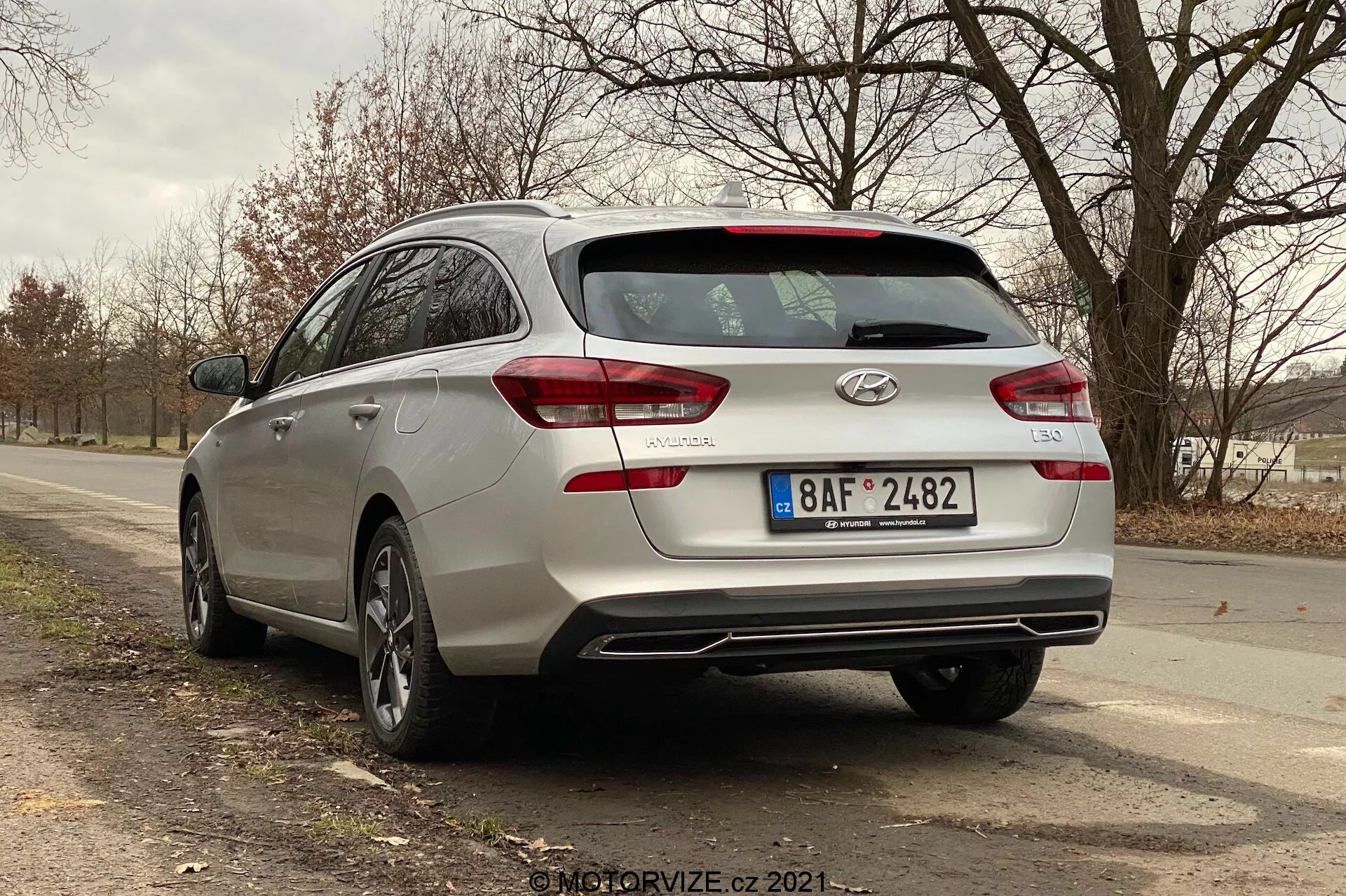 Vista tres cuartos trasera de un Hyundai i30 familiar plateado del 2020 que muestra los elementos de diseño trasero, incluidas las luces traseras posiblemente con tecnología LED, limpiaparabrisas trasero, insignias y nombre del modelo del coche. La imagen también captura las dos salidas de escape integradas en el parachoques trasero, llantas de aleación y un perfil lateral parcial que sugiere un amplio espacio en la cabina. El coche está estacionado en una carretera de dos carriles en una zona rural o semirrural con árboles y espacio abierto al fondo.