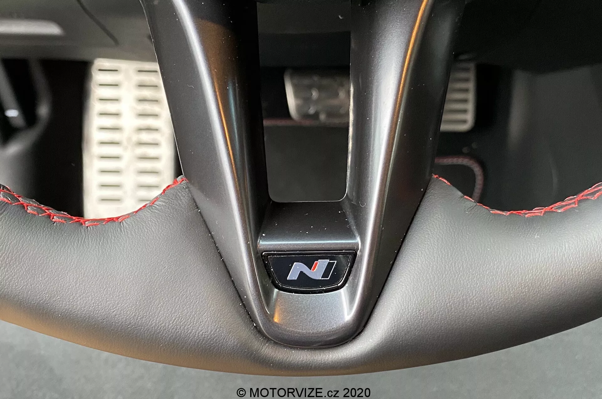 Vista interior en primer plano del volante del Hyundai i30 2020 (i30-kombi) desde abajo, destacando la sección inferior con un emblema central 'N' indicativo de una variante de alto rendimiento, acabado en cuero con costuras rojas, radios metálicos y detalles de pedales deportivos perforados en el fondo desenfocado.