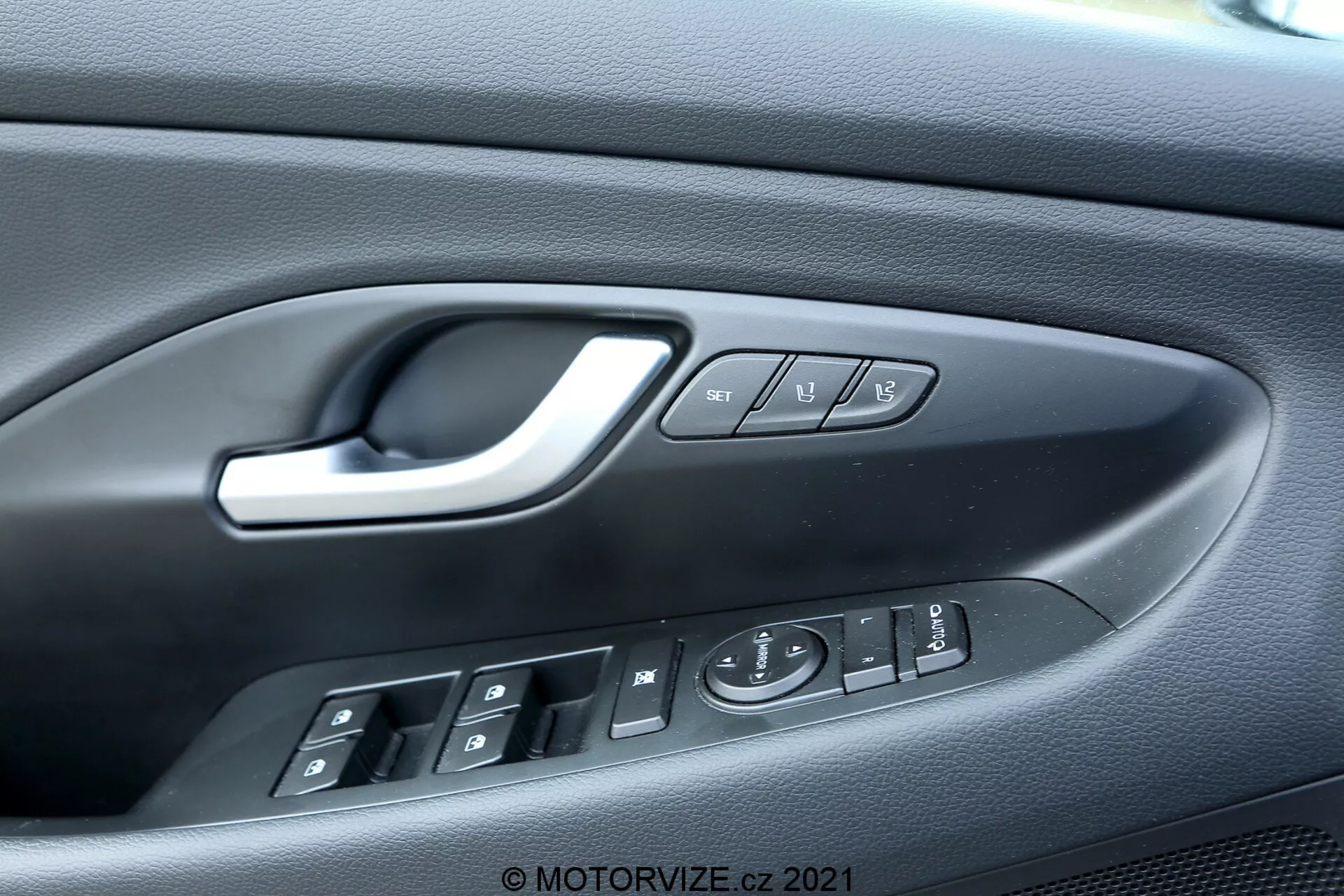 Vista en primer plano del panel de la puerta del lado del conductor del Hyundai i30 Kombi 2020, destacando el diseño intrincado y la funcionalidad que incluye el tirador de puerta ergonómico plateado, cuatro botones de control de ventanas eléctricas con indicación de bajada automática, un joystick electrónico para ajustes de los espejos laterales, botones de bloqueo y desbloqueo, y botones de memoria del asiento. El panel de plástico texturizado agrega estética y agarre, indicando un interior con muchas funciones.