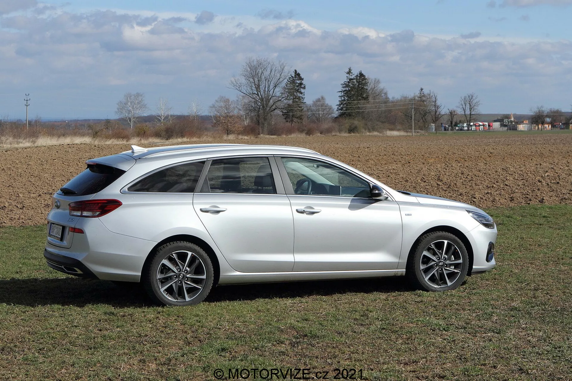 Vista lateral del Hyundai i30 Kombi 2020 en un campo de hierba a la luz del día. El coche tiene llantas de aleación multirradio, raíles en el techo y ventanas traseras tintadas, situado en un entorno rural con campos agrícolas y un cielo parcialmente nublado al fondo.