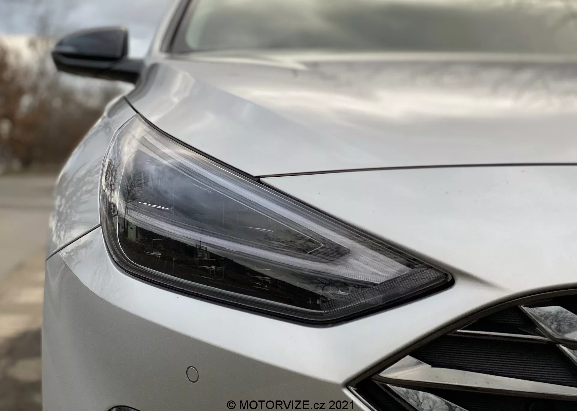 Toma en primer plano del Hyundai i30 2020 Kombi, mostrando la sección delantera izquierda del vehículo en un ángulo de tres cuartos hacia el lado del conductor, enfocándose en el conjunto de faros elegantes y angulares con tecnología LED o HID moderna, acompañado por un espejo lateral aerodinámico con posibles integraciones tecnológicas avanzadas como ajuste eléctrico y monitoreo de puntos ciegos. La carrocería del coche presenta un acabado perlado o metálico de alta calidad con contornos aerodinámicos en el capó y una pista de la rejilla delantera. El fondo está desenfocado para enfatizar los detalles del coche.