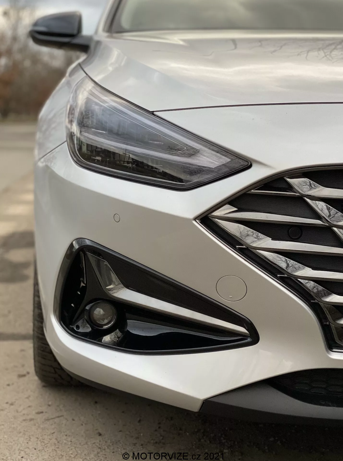 Fotografía angular de cerca de la parte delantera izquierda de un moderno Hyundai i30 Kombi 2020, enfocándose en el faro y el área del parachoques delantero. Tomada desde un ángulo bajo cerca de la rueda delantera, la imagen captura el faro LED angular, la parrilla con detalles cromados, el diseño agresivo del parachoques con una gran entrada de aire y la luz antiniebla incrustada en el adorno negro. El acabado metálico de la pintura refleja la luz, y las líneas de diseño en el capó enfatizan el aspecto dinámico del coche. El fondo está desenfocado para resaltar los detalles del vehículo, sugiriendo funciones avanzadas como iluminación adaptativa y posibles sensores de estacionamiento.