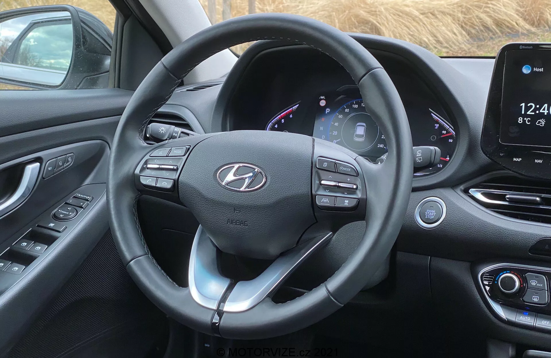 Imagen del interior de la cabina delantera del Hyundai i30 2020 desde la perspectiva del conductor. El enfoque está en el volante moderno con múltiples botones de control y paletas de cambio, forro de cuero con costuras visibles y un emblema de logotipo centrado. Detrás del volante hay un cuadro de instrumentos parcialmente digital que muestra el velocímetro, tacómetro, indicador de combustible y otra información del vehículo con una interfaz colorida. A la derecha, la consola central presenta un sistema de infoentretenimiento con pantalla táctil que muestra la hora, la temperatura y la interfaz de inicio, con botones y diales de control climático físicos debajo. La cabina cuenta con un diseño contemporáneo con superficies curvas, materiales suaves al tacto y un esquema de color negro con acentos plateados o cromados que realzan la apariencia moderna y sofisticada.