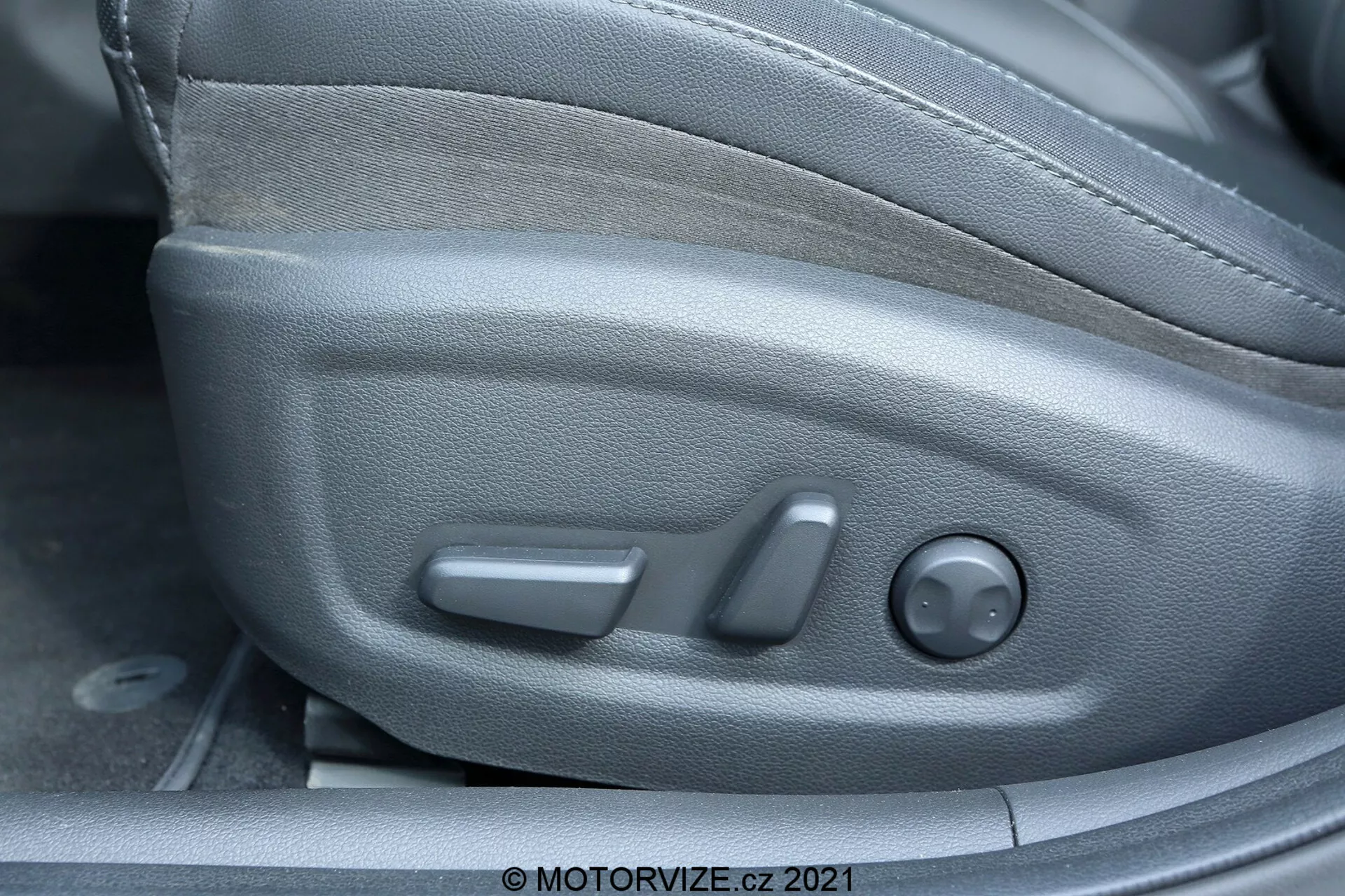 Vista de cerca del interior del Hyundai i30 2020, centrándose en los controles del asiento del conductor desde un ángulo lateral. Las características visibles incluyen controles de ajuste del asiento: una barra delantera para deslizar, una palanca central para reclinar y un control redondo en la parte trasera para ajustar la altura. La cubierta de plástico se integra con el estilo lateral del asiento, y la tapicería de tela ofrece durabilidad y comodidad.
